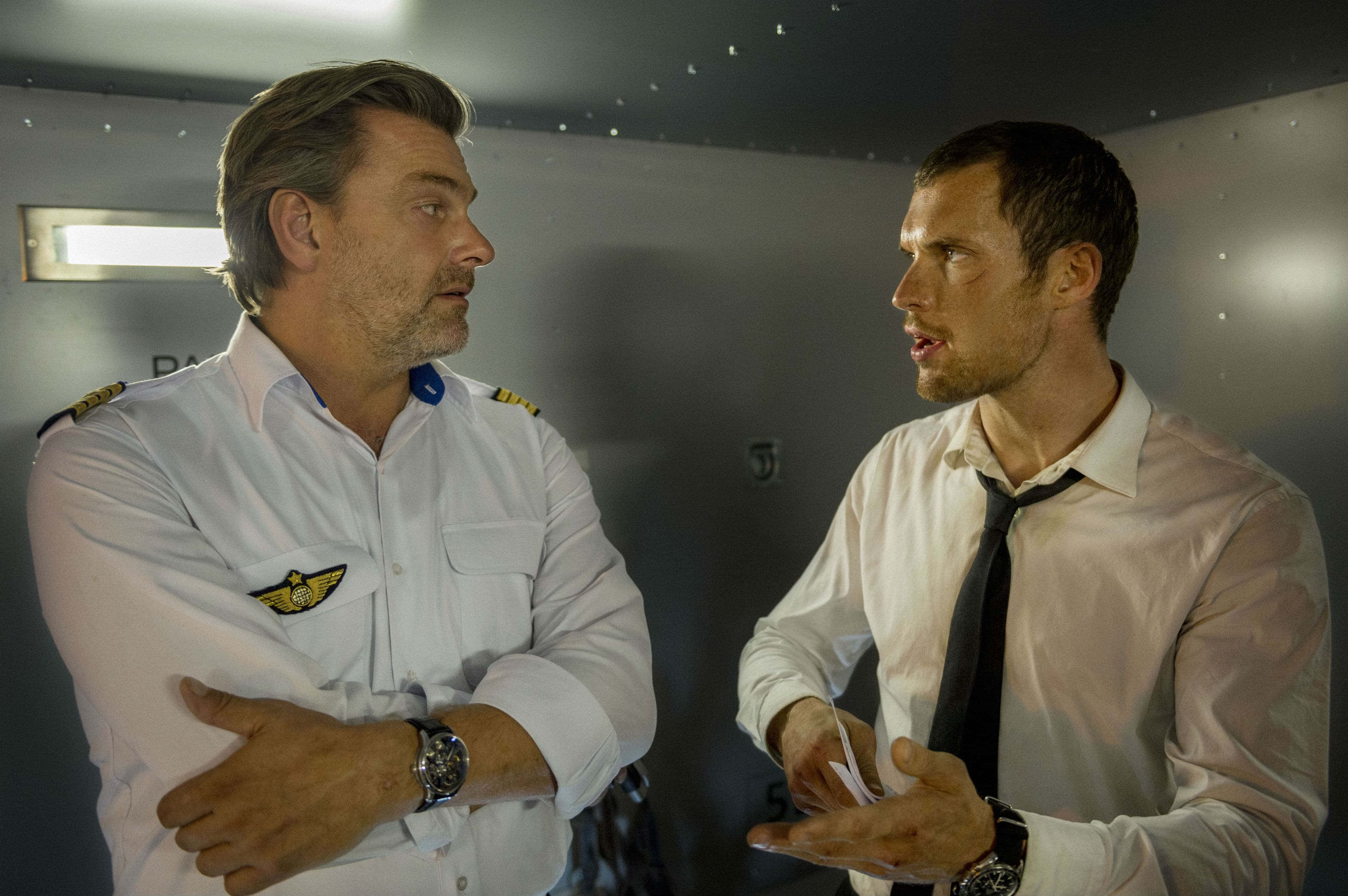 Ray Stevenson 桌布 壁紙 — 兩名身穿機師和商務服裝的男子在燈光昏暗的飛機艙內進行緊張對話。