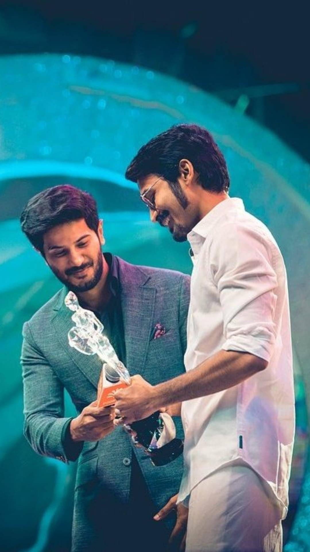 Dhanush 桌布 壁紙 — 兩名身穿正裝的男子在充滿活力的藍色舞台燈光下交換水晶獎項。