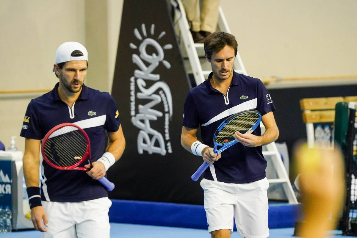 Tennis 桌布 壁紙 — 兩名身穿海軍藍和白色制服的男網球選手在藍色室內球場上手持球拍行走。
