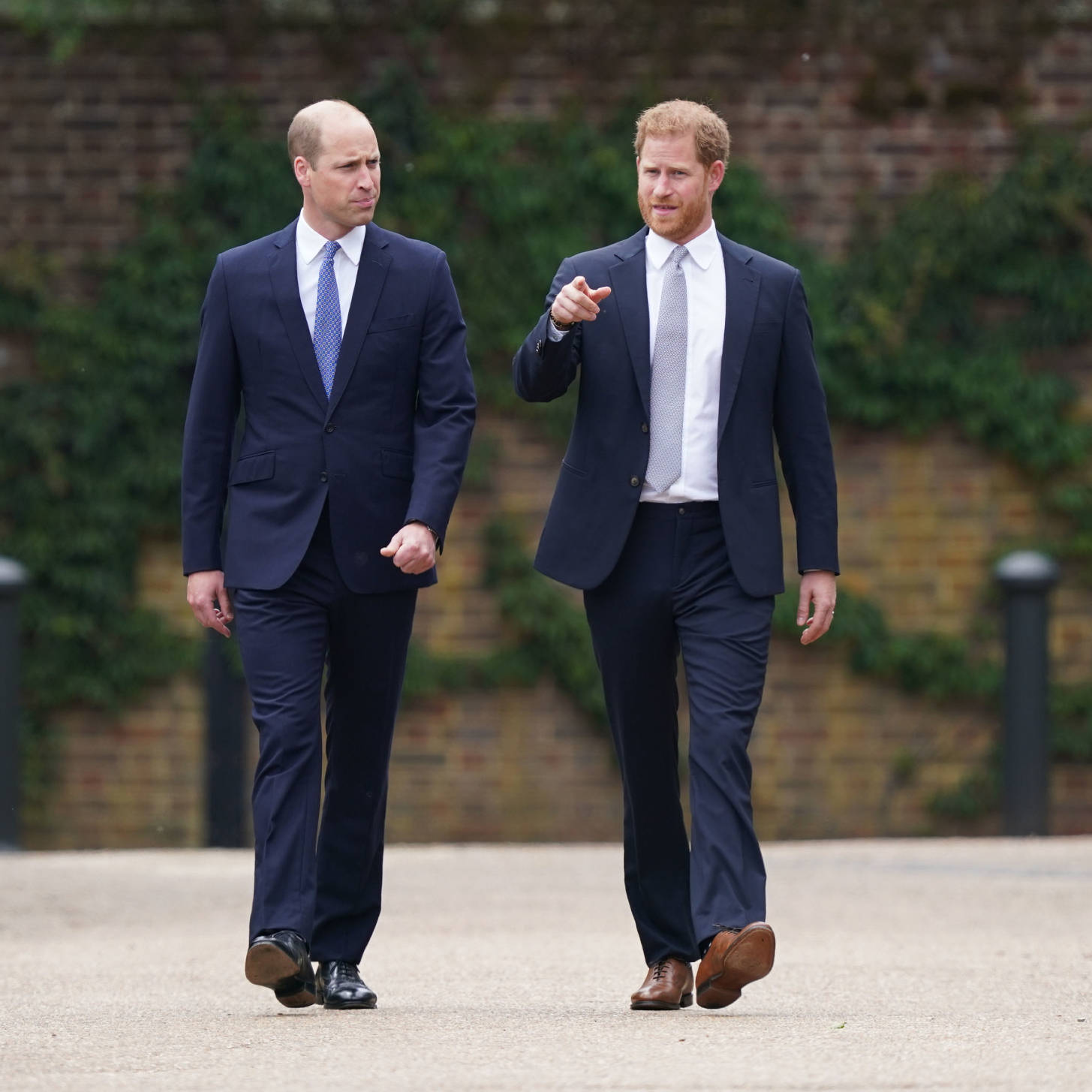 Royal 桌布 壁紙 — 兩名身穿海軍藍西裝的男子並肩走在鋪砌的小徑上，身後有磚牆和綠植。