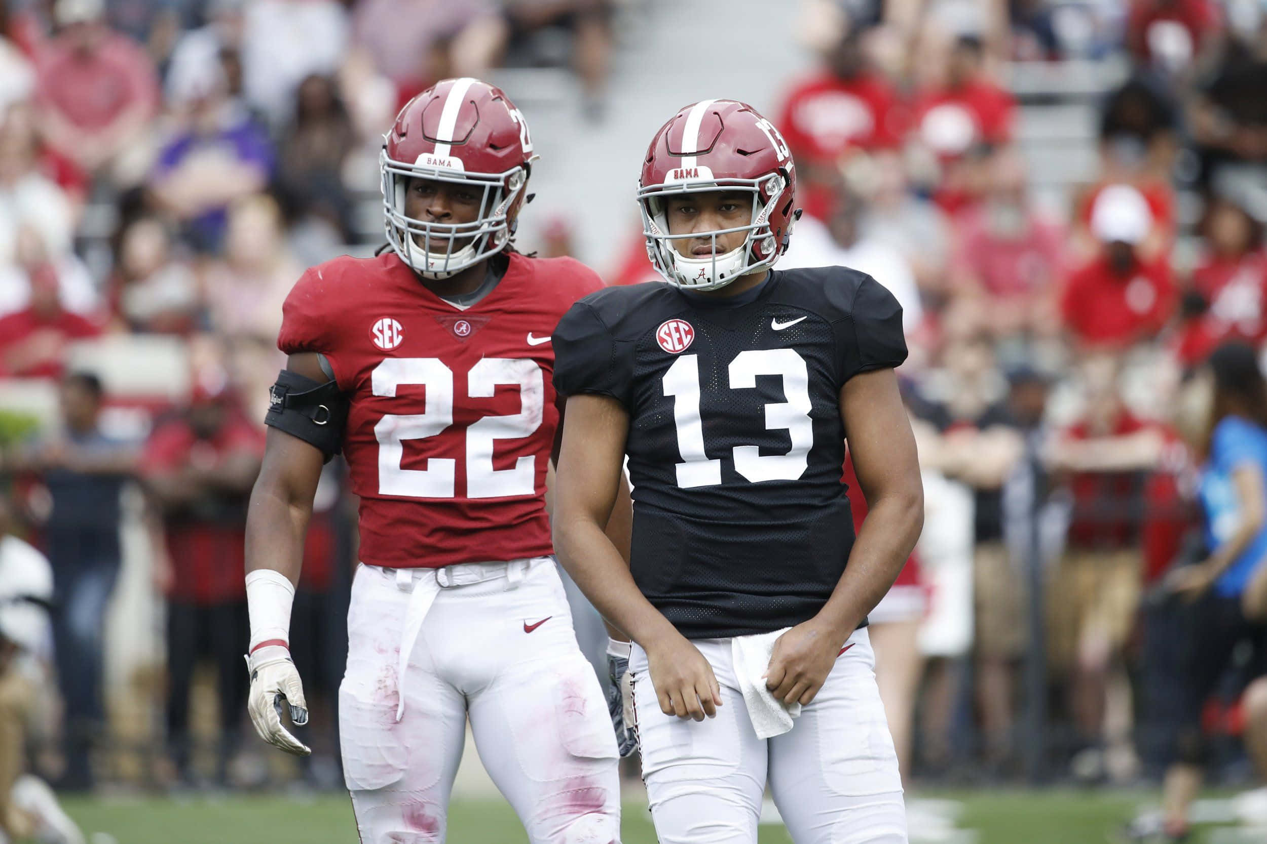 Alabama Football 桌布 壁紙 — 兩名身穿深紅和黑色制服的美式足球員，穿著沾滿泥濘的褲子和頭盔，在明亮的日光下站在球場上。