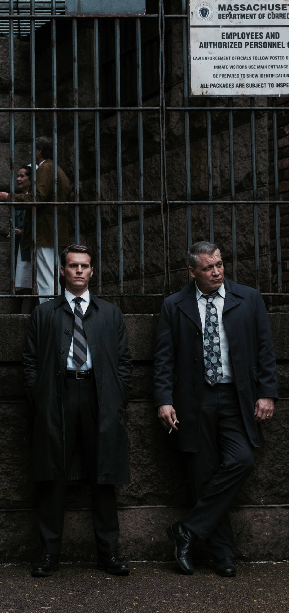 Netflix 桌布 壁紙 — 兩名身穿深色外套的男子站在麻薩諸塞州監獄外的石牆旁，監獄設有鐵欄杆大門和標誌。