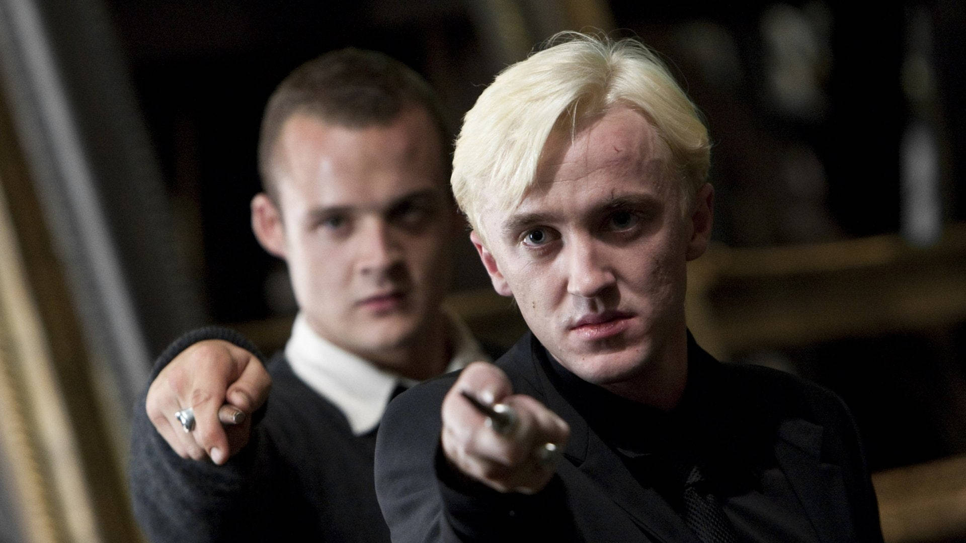 draco 桌布 壁紙 — 兩名身穿深色衣服的男子在昏暗的室內環境中，帶著強烈的表情向前指。