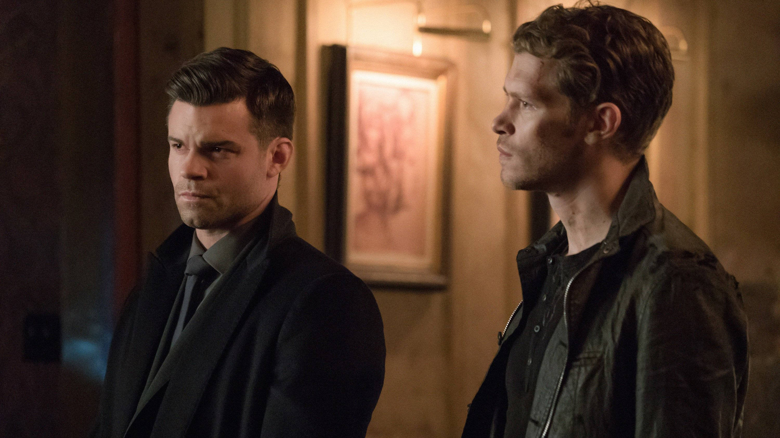 elijah mikaelson 桌布 壁紙 — 兩名身穿深色衣服的男子站在一個光線溫暖的室內走廊，身後牆上掛著一幅帶框的畫作。