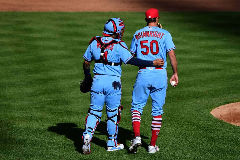 Yadier Molina 桌布 壁紙 — 兩名身穿淺藍色球衣的棒球運動員在陽光明媚的綠色球場上一起行走，一人穿著捕手裝備，另一人拿著棒球。