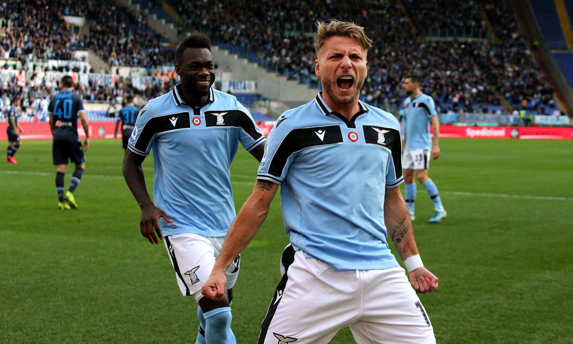 ciro immobile 桌布 壁紙 — 兩名身穿淺藍色球衣的足球運動員在充滿活力的綠色球場上，在擠滿觀眾的體育場前熱情慶祝。