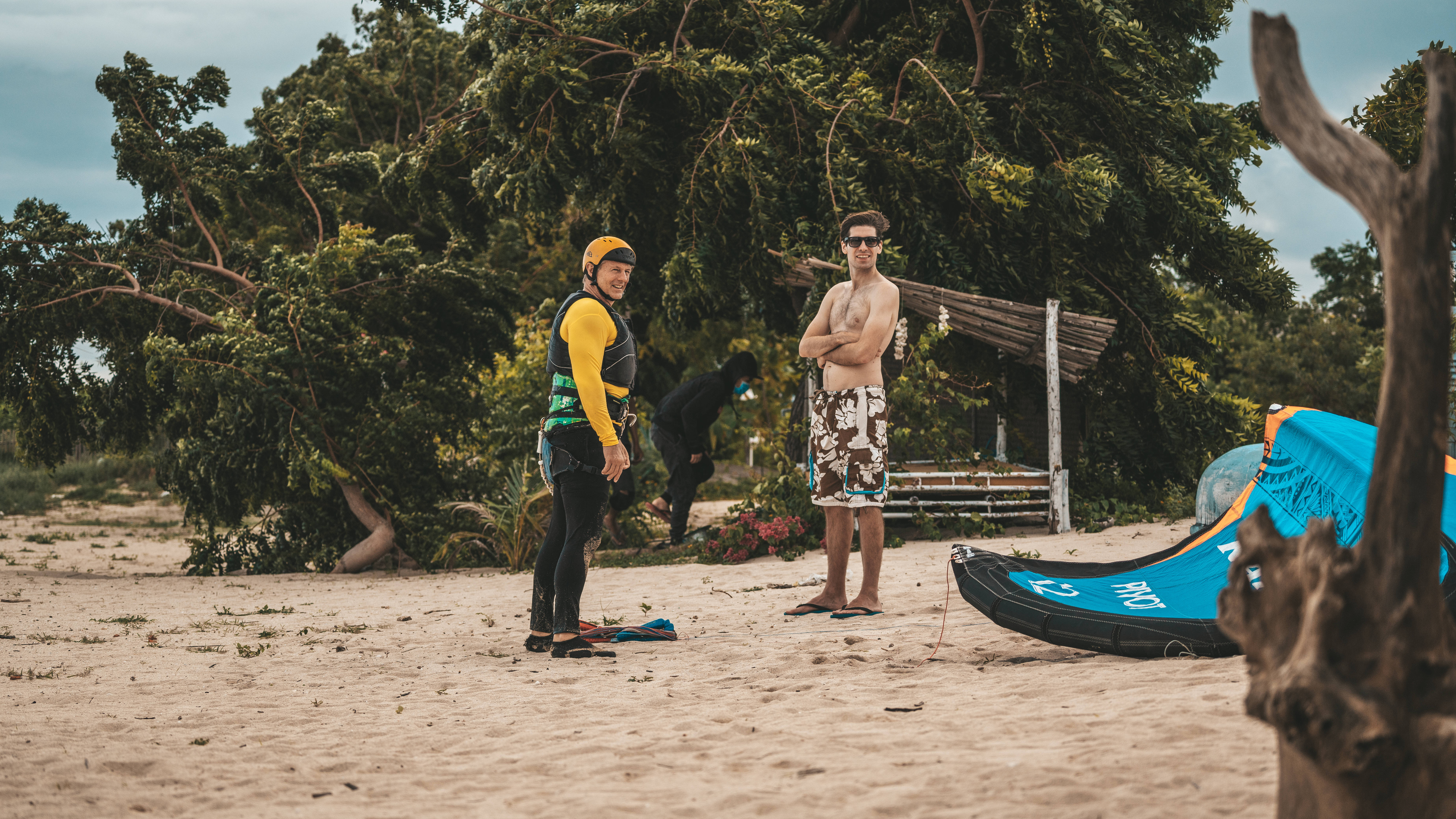 Windsurfing 桌布 壁紙 — 兩名身穿潛水衣和太陽眼鏡的男子站在沙灘上，旁邊有一個藍色風箏板和一棵大樹，背景有一個木製遮蔽處。