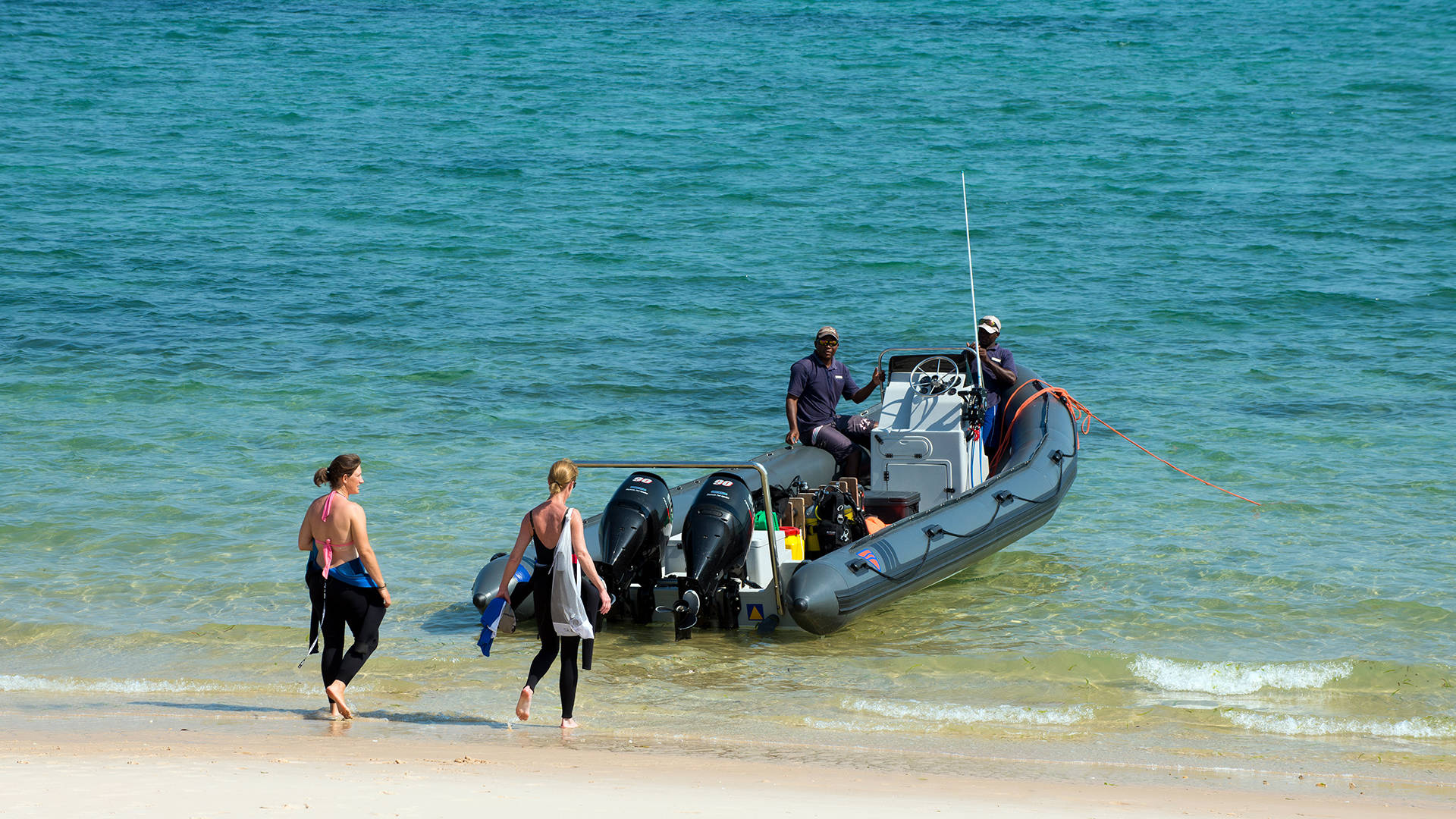 Mozambique 桌布 壁紙 — 兩名身穿潛水衣的女子在陽光明媚、海水碧綠的海灘上，走向一艘載有兩名男子的灰色充氣艇。