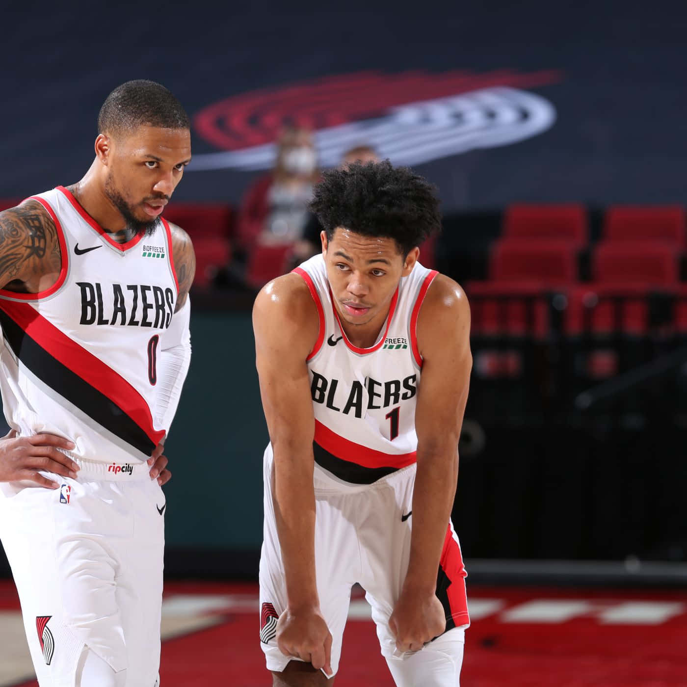 Anfernee Simons 桌布 壁紙 — 兩名身穿白色、紅色和黑色球衣的波特蘭拓荒者籃球運動員在比賽中專注地站在球場上，背景有紅色座位和球隊標誌。