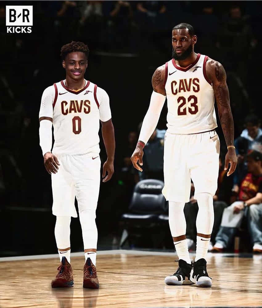 Bronny James 桌布 壁紙 — 兩名身穿白色Cavs球衣的籃球運動員站在拋光球場上，場館燈光下，身後有模糊的觀眾。