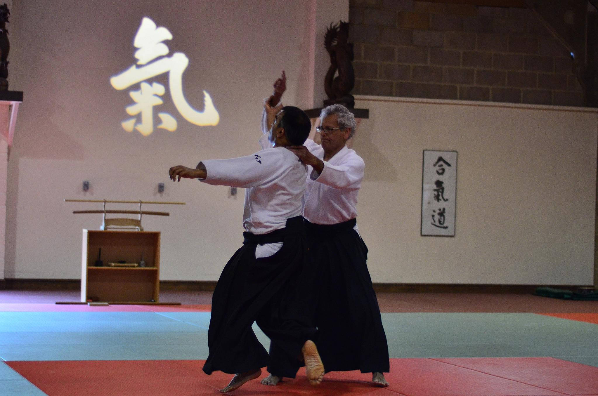martial arts 桌布 壁紙 — 兩名身穿白色上衣和黑色袴的合氣道練習者在發光漢字投影下的紅藍色墊子上進行動態投技。