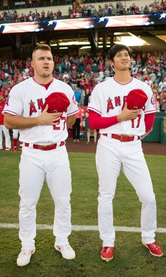 Mike Trout 桌布 壁紙 — 兩名身穿白色天使隊球衣的棒球運動員站在綠色球場上，在國歌奏起時將紅色帽子放在心口。