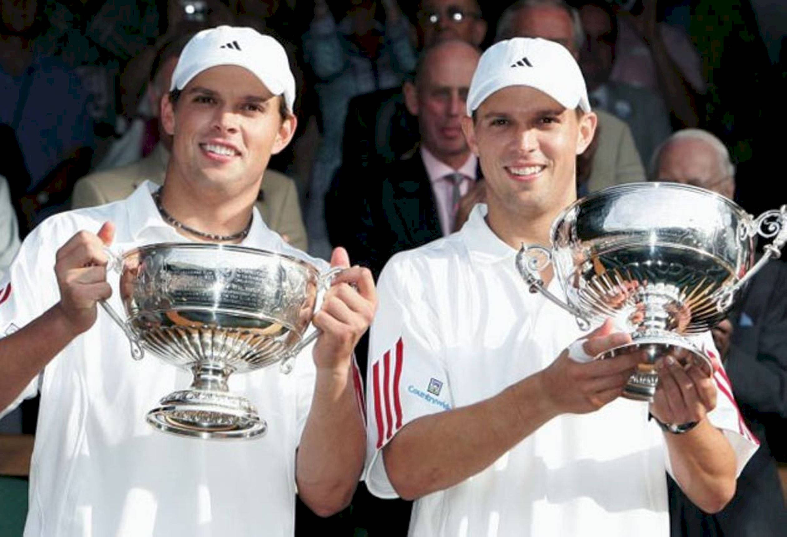 Trophy 桌布 壁紙 — 兩名身穿白色球衣、面帶笑容的男性網球選手，在陽光明媚的戶外活動中自豪地高舉大型銀色獎盃，身後有觀眾。