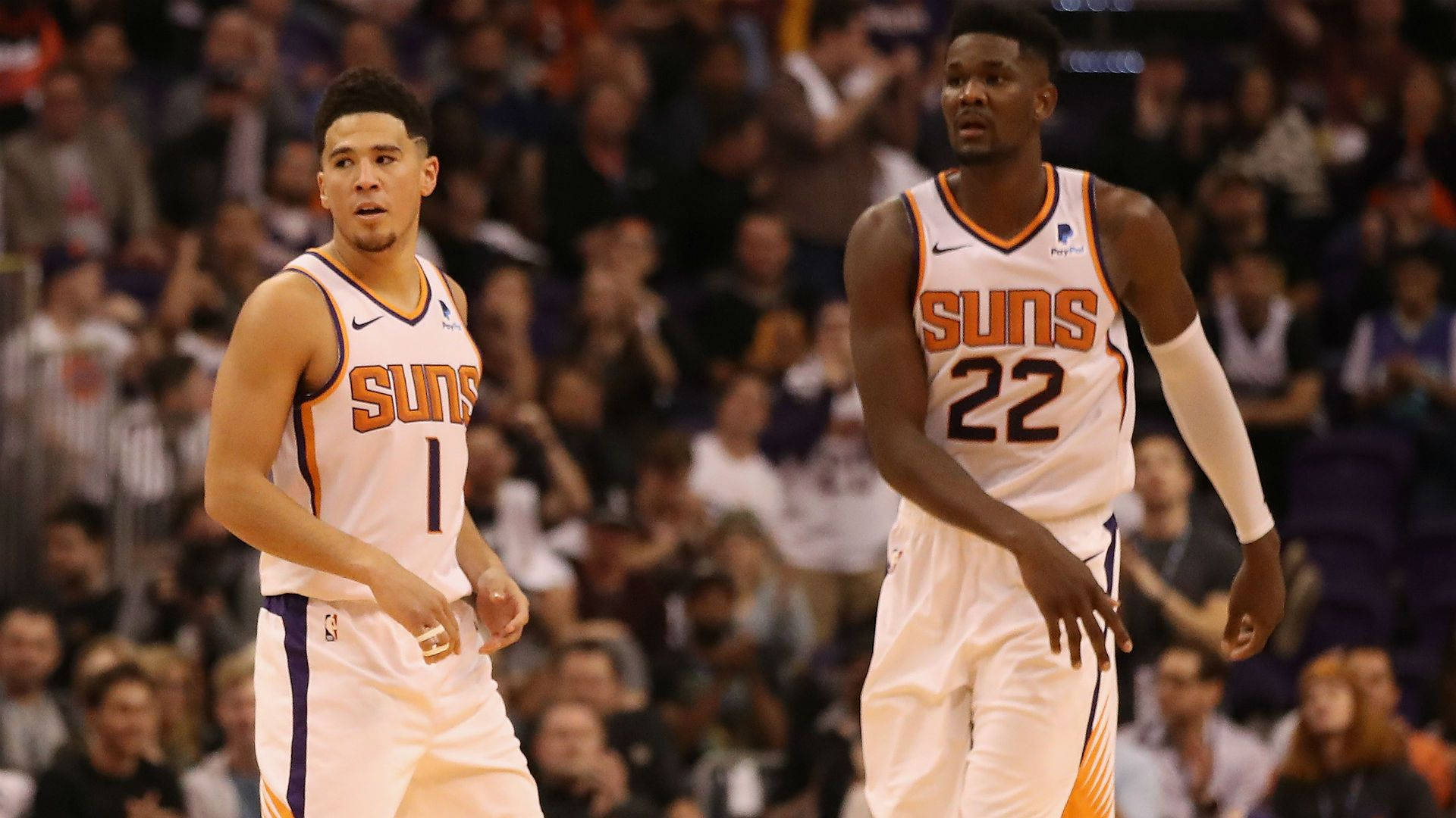 Devin Booker 桌布 壁紙 — 兩名身穿白色球衣的鳳凰城太陽隊籃球運動員在比賽中專注地站在球場上，身後是模糊的人群。