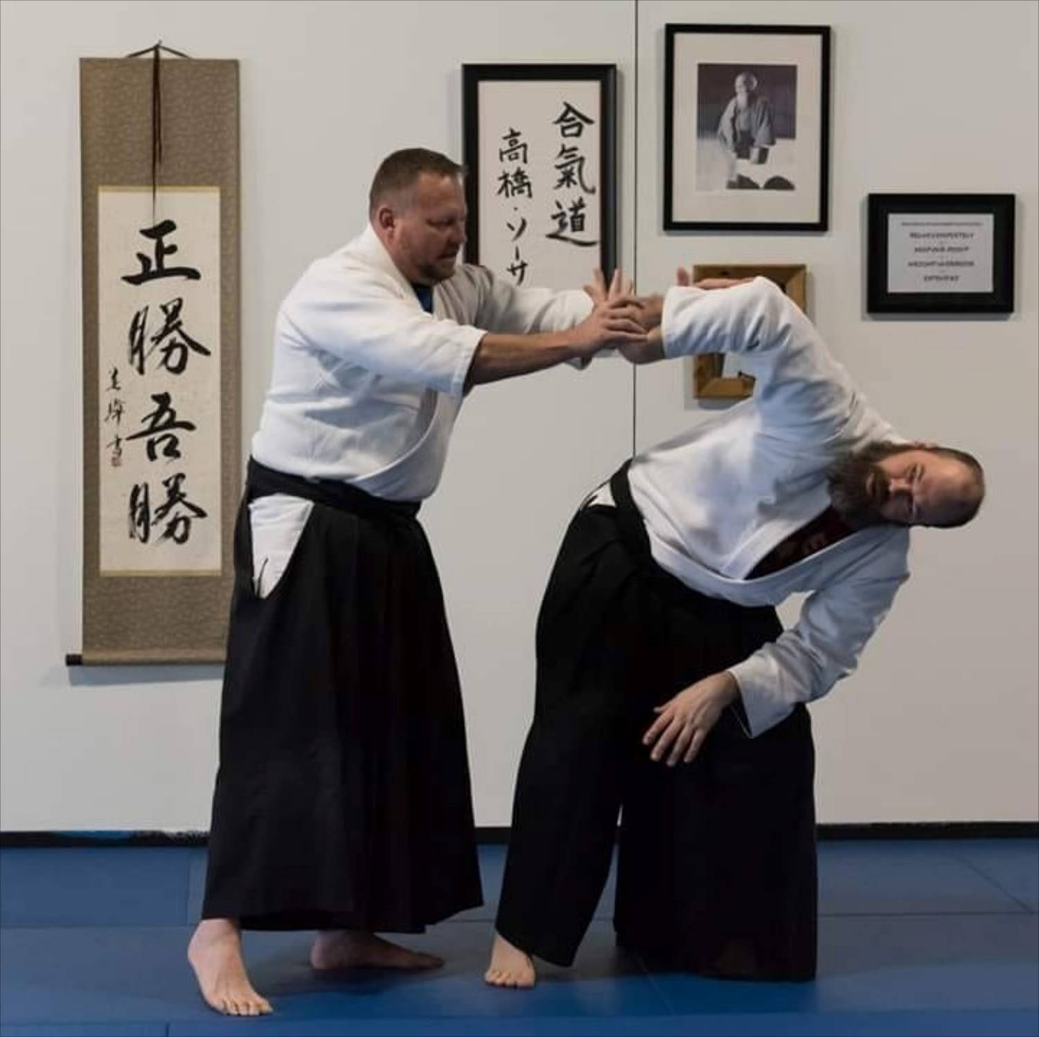 martial arts 桌布 壁紙 — 兩名身穿白色道服和黑色袴的男子在藍色軟墊上練習合氣道投技，牆上掛有書法卷軸。