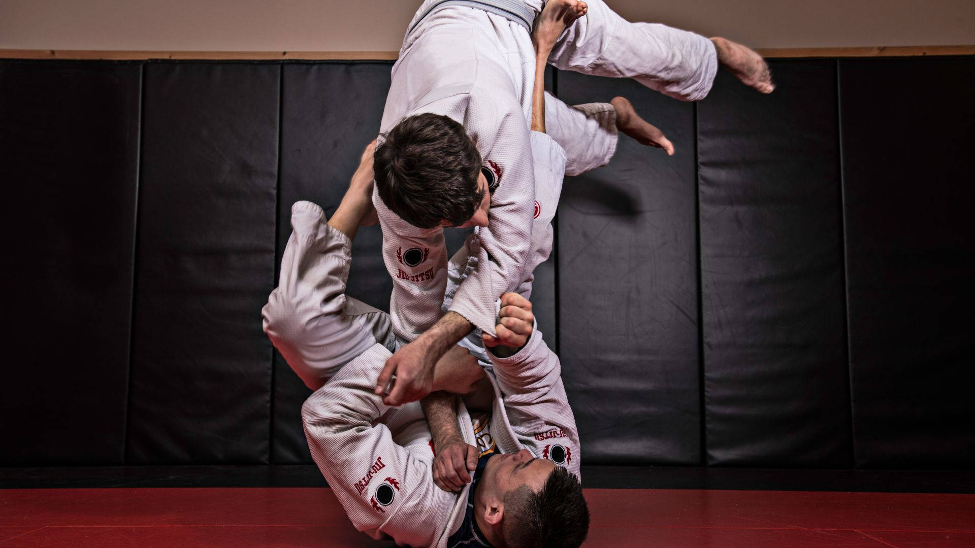 martial arts 桌布 壁紙 — 兩名身穿白色道服的武術家在黑色軟墊牆壁前的紅色軟墊上執行一個動態的纏鬥動作。