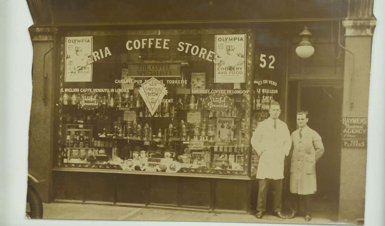 Old Times 桌布 壁紙 — 兩名身穿白袍的男子自豪地站在一間復古咖啡店外，店內玻璃陳列櫃擺滿了罐子和宣傳Olympia咖啡的標誌。