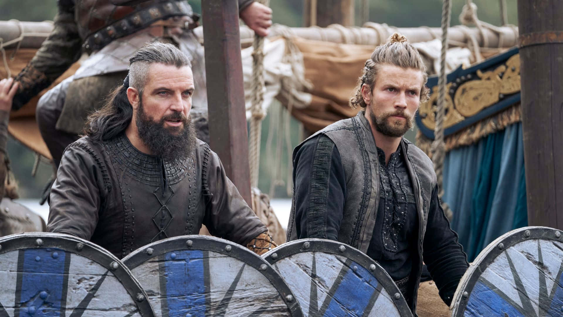 Vikings 桌布 壁紙 — 兩名身穿皮革盔甲、留鬍鬚的維京戰士站在木製長船上，手持帶有藍白圖案的圓盾。