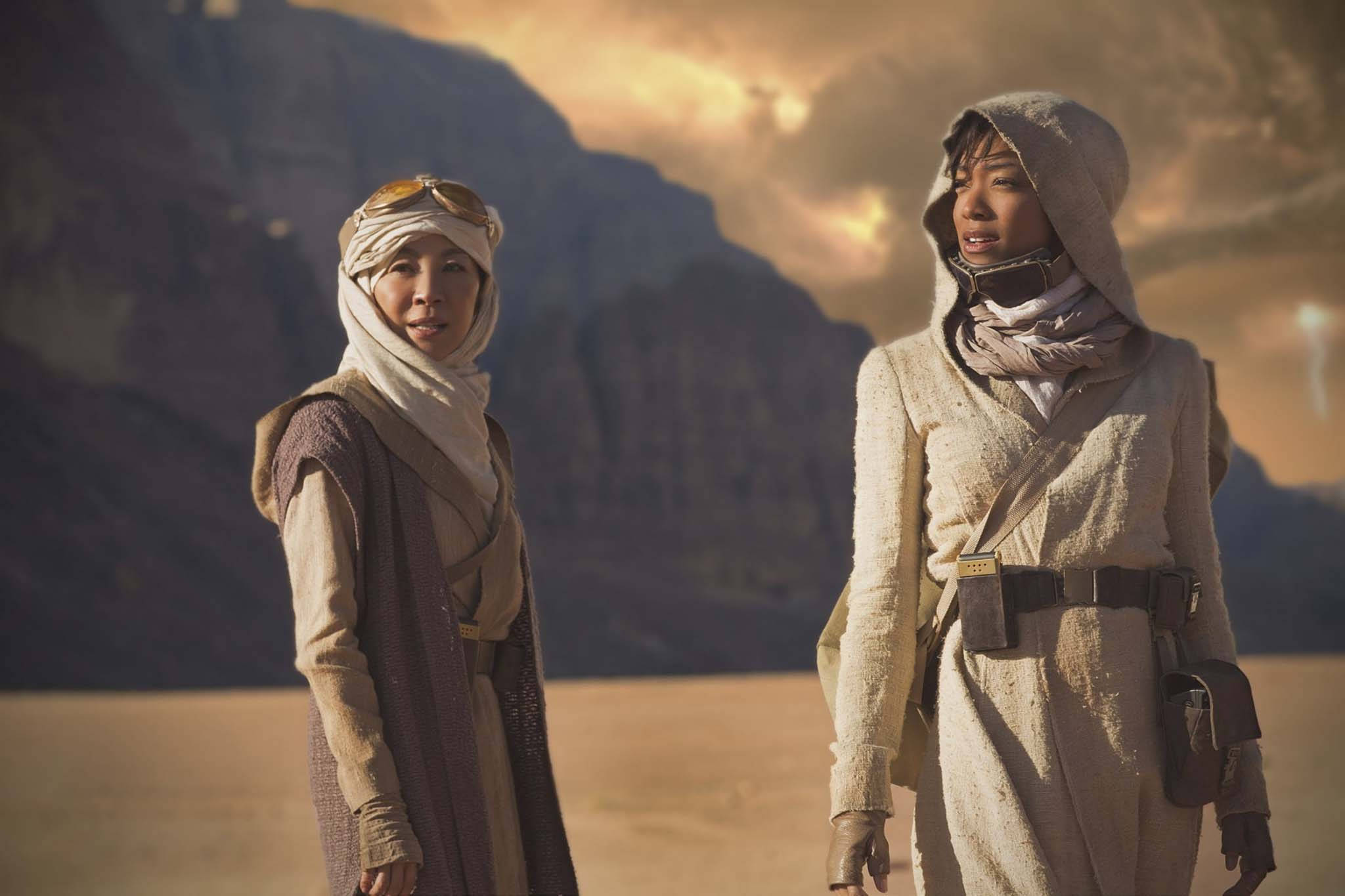 Discovery 桌布 壁紙 — 兩名身穿破舊長袍的沙漠旅人，在金色夕陽天空下，以崎嶇山脈為背景站立。