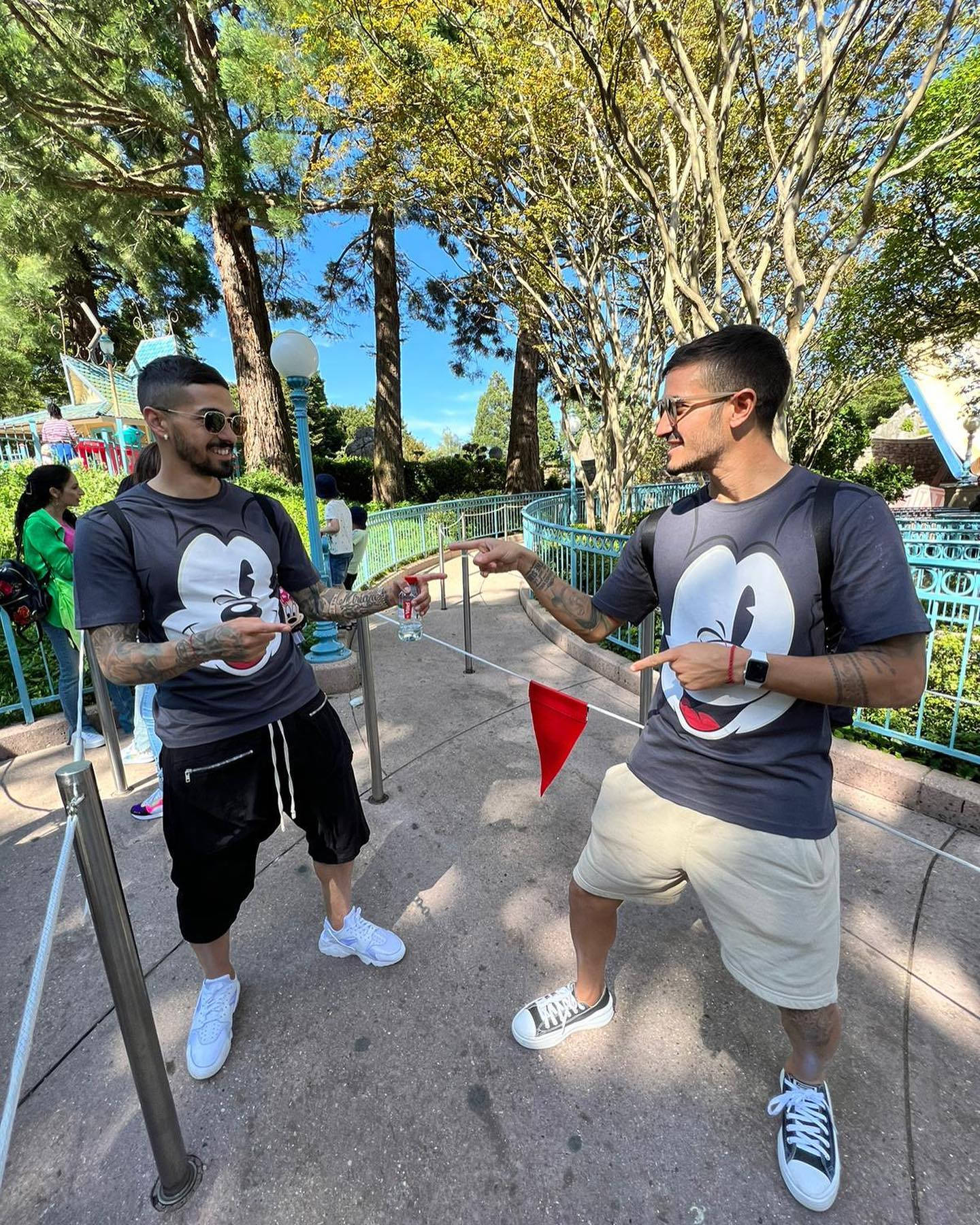 Football Players 桌布 壁紙 — 兩名身穿米奇老鼠T恤的男子在主題公園陽光普照的樹下，以指向手勢俏皮地擺姿勢。