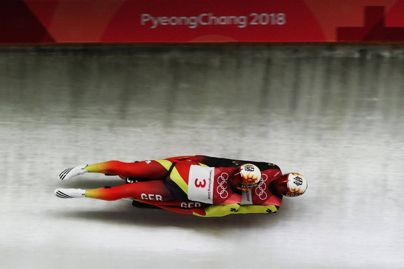 luge 桌布 壁紙 — 兩名身穿紅、黃、黑色雪橇服的運動員在平昌2018的冰道上頭朝下競速。