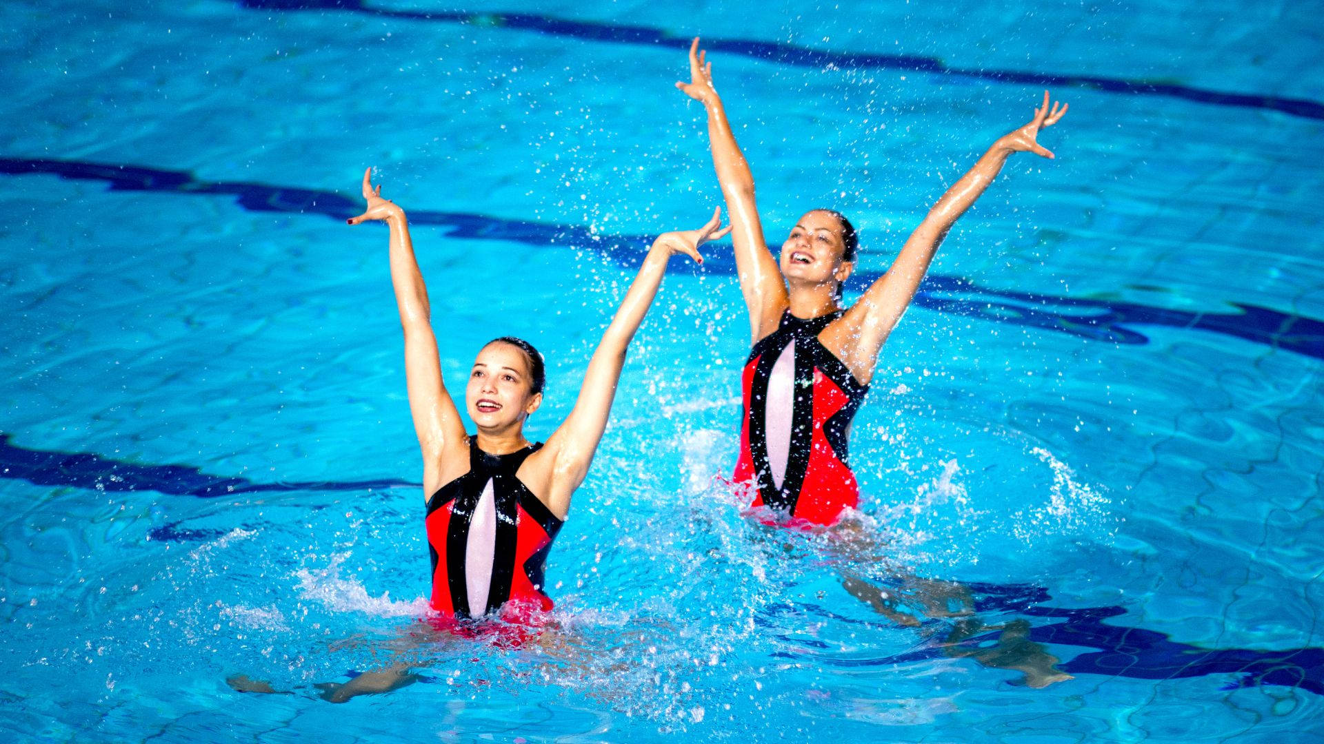 Swimming 桌布 壁紙 — 兩名身穿紅、黑、白色服裝的韻律泳選手在充滿活力的藍色泳池水中舉臂歡樂濺水。