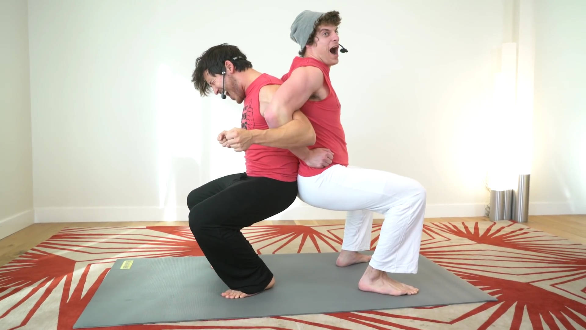 Markiplier 桌布 壁紙 — 兩名身穿紅色上衣的男子在明亮的房間內，於紅色圖案地毯上的灰色墊子上做出動態瑜伽姿勢。