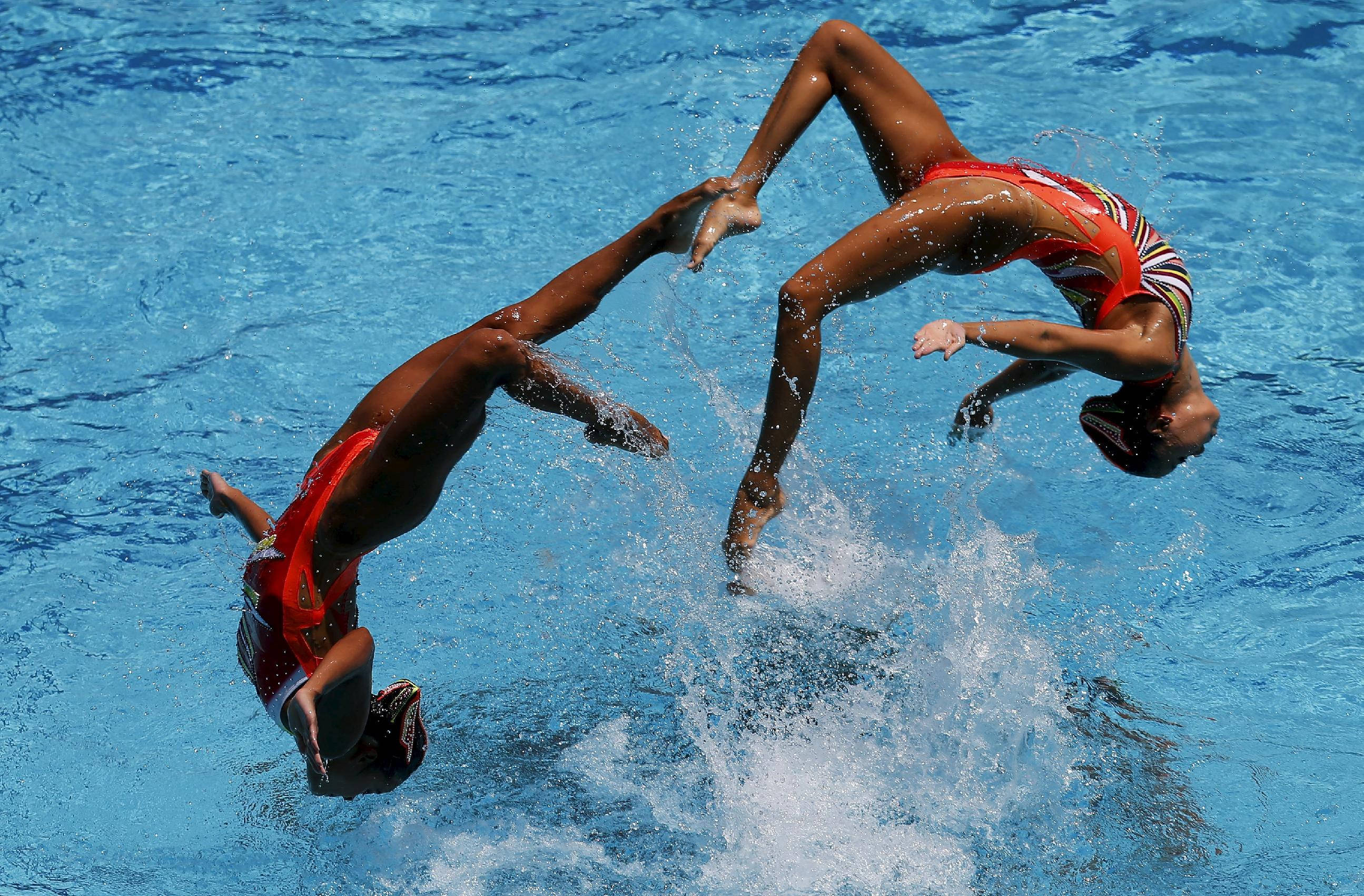 Swimming 桌布 壁紙 — 兩名身穿紅色和白色泳衣的韻律泳運動員在明亮的藍色水中執行動態的頭頂分腿動作。