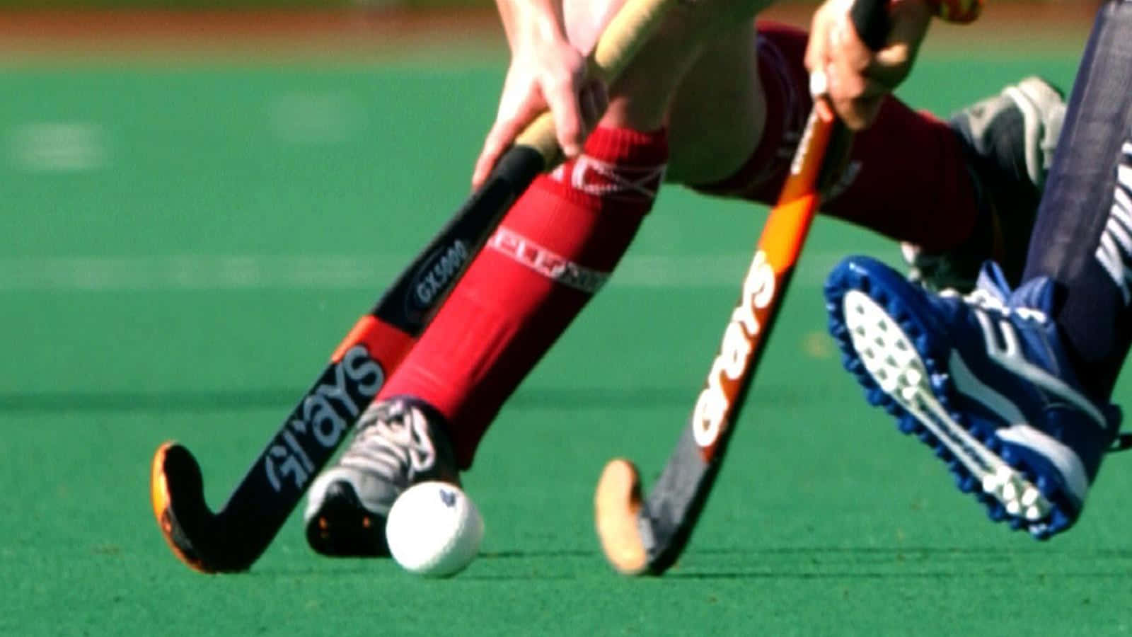 hockey 桌布 壁紙 — 兩名身穿紅色和藍色裝備的曲棍球運動員在充滿活力的綠色草地上激烈爭奪控球權。