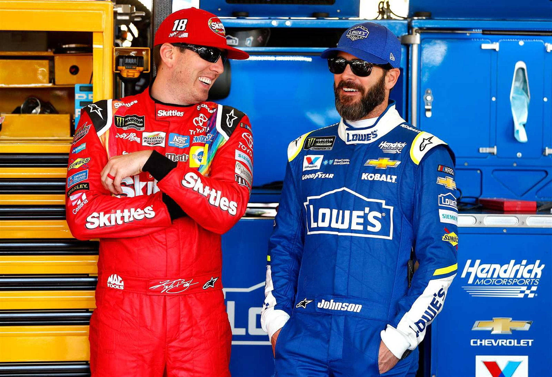 Racing 桌布 壁紙 — 兩名身穿紅色和藍色賽車服的NASCAR車手在一個有黃色工具箱和藍色設備的車庫裡一起大笑。