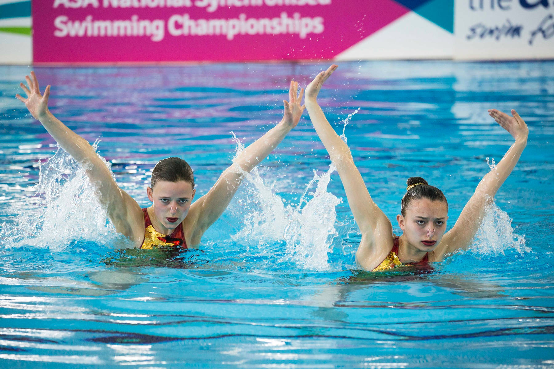 Swimming 桌布 壁紙 — 兩名身穿紅色和金色泳衣的韻律泳選手在錦標賽中做出雙臂舉出水面的動態動作。