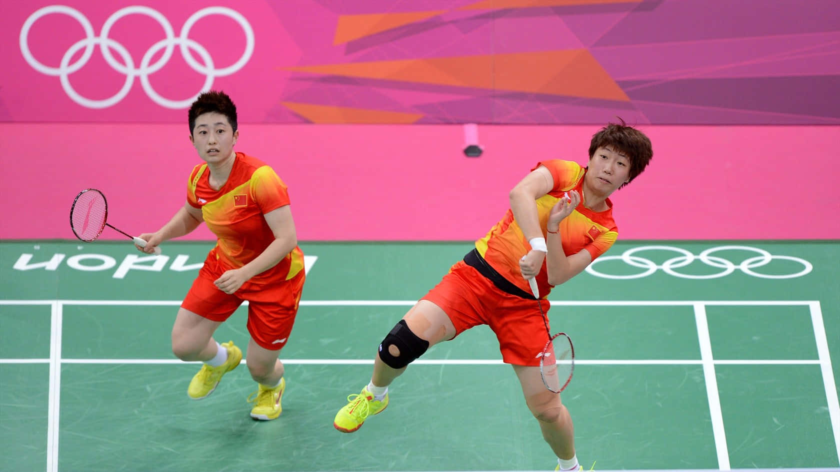 HD Badminton 桌布 壁紙 — 兩名身穿紅色和黃色球衣的羽球選手在明亮的場館燈光下於綠色球場上激烈競爭。