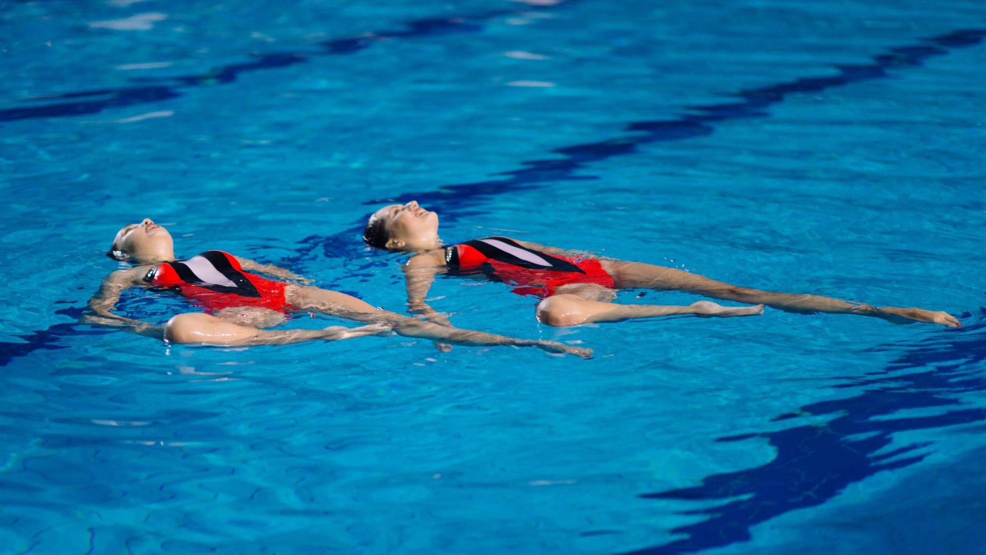 Swimming 桌布 壁紙 — 兩名身穿紅色和黑色泳衣的韻律泳選手在充滿活力的藍色泳池水中優雅地仰臥漂浮。