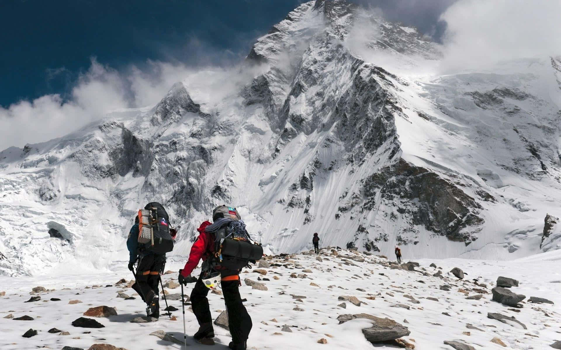 Hiking 桌布 壁紙 — 兩名身穿紅色和黑色裝備的登山者穿越白雪覆蓋的山脊，身後聳立著雲霧繚繞的高聳山峰。