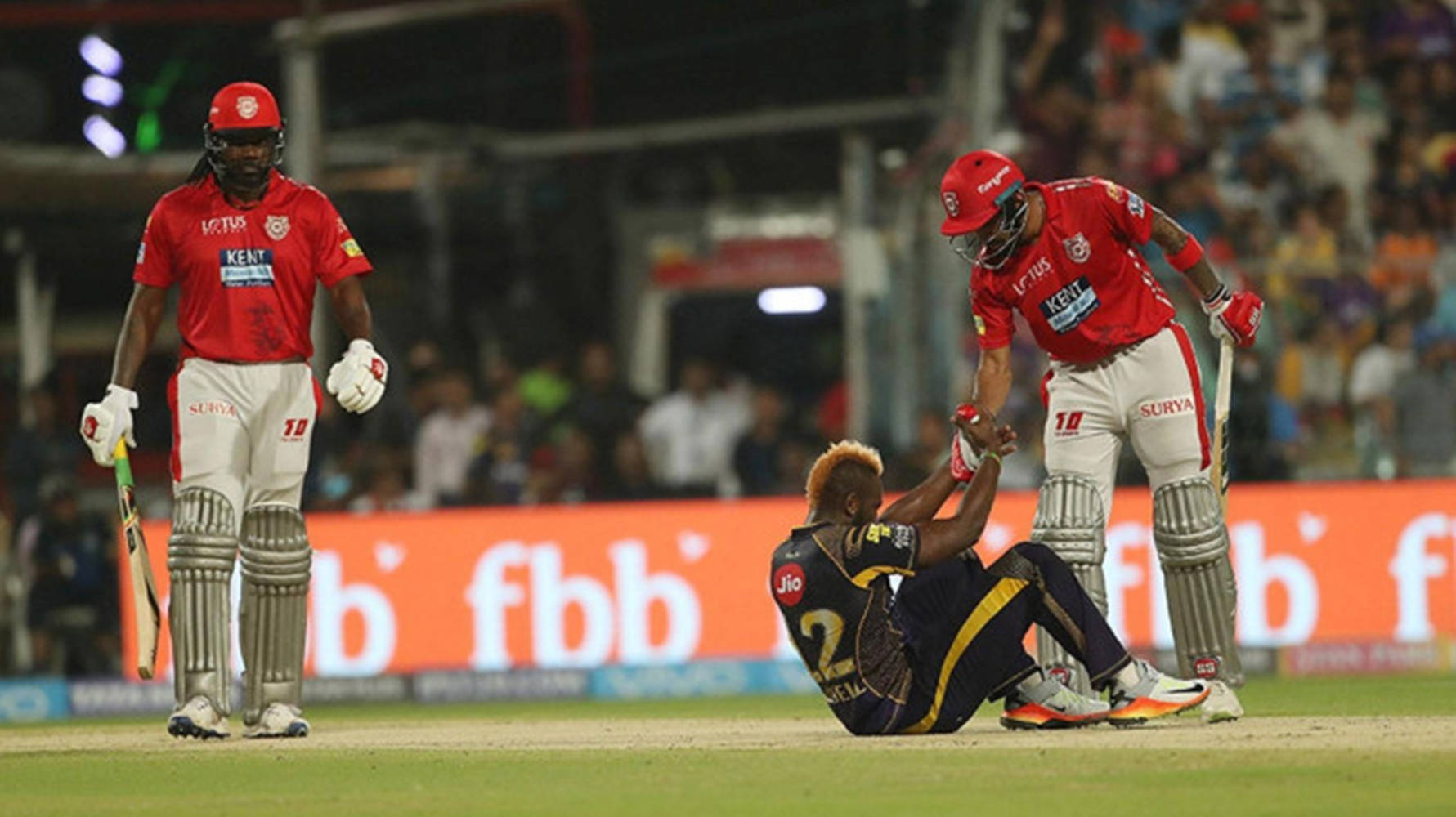 kl rahul 桌布 壁紙 — 兩名身穿紅色球衣的板球運動員在燈光明亮的體育場場地上，協助一名身穿海軍藍裝備的倒地隊友。