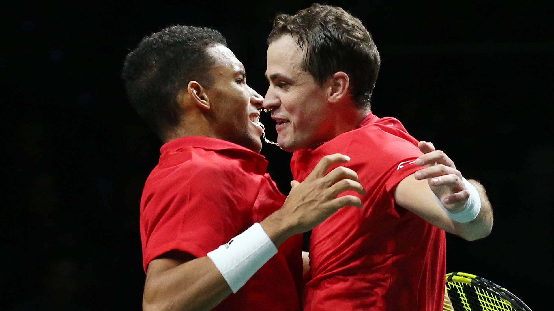 felix auger aliassime 桌布 壁紙 — 兩名身穿紅色球衣的男網球選手在明亮的體育場燈光下，賽後愉快地擁抱。