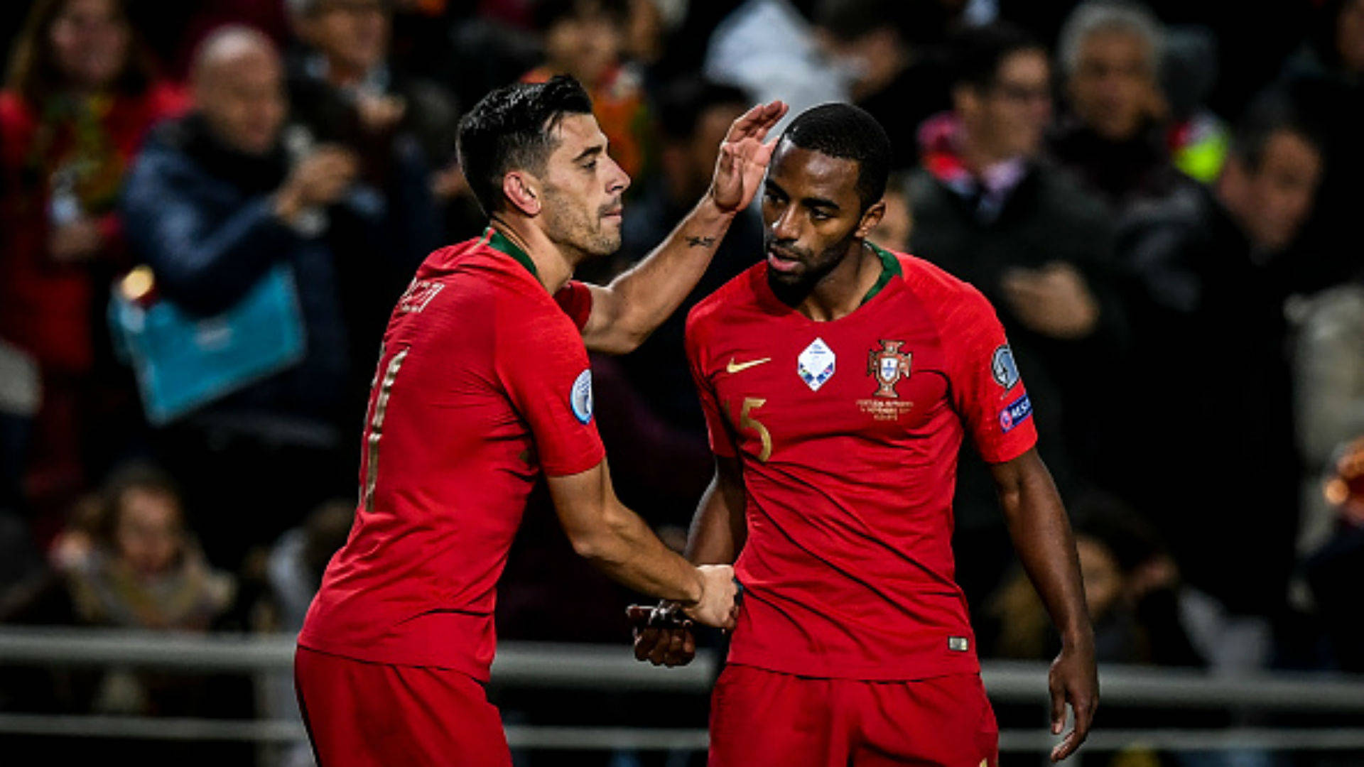 Football Players 桌布 壁紙 — 兩名身穿紅色球衣的葡萄牙足球員在球場燈光下以握手和輕拍頭部慶祝進球。