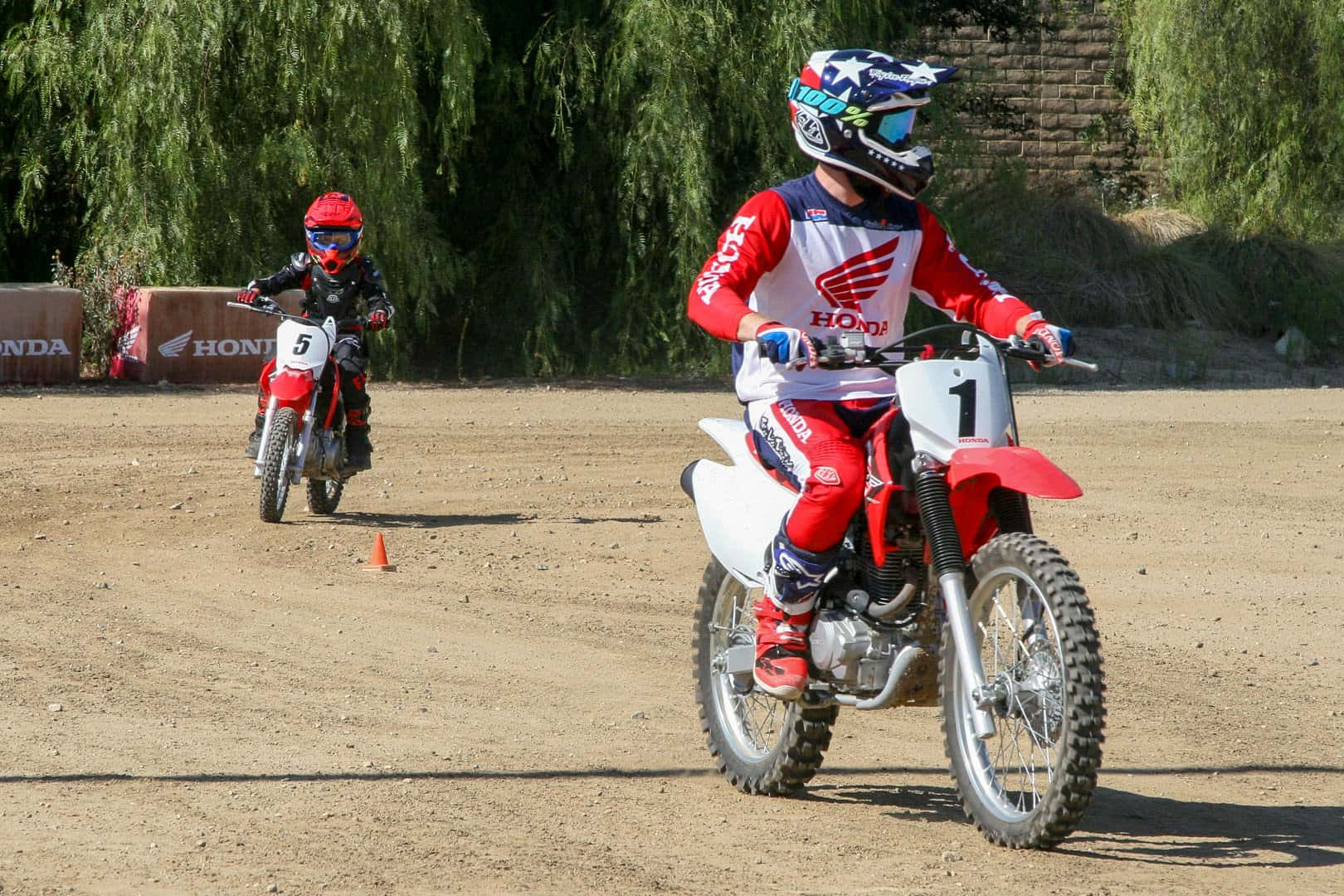 Dirtbike 桌布 壁紙 — 兩名身穿紅色裝備的年輕越野摩托車手，在陽光明媚、塵土飛揚的賽道上駕駛紅色本田越野摩托車，背景有綠樹。