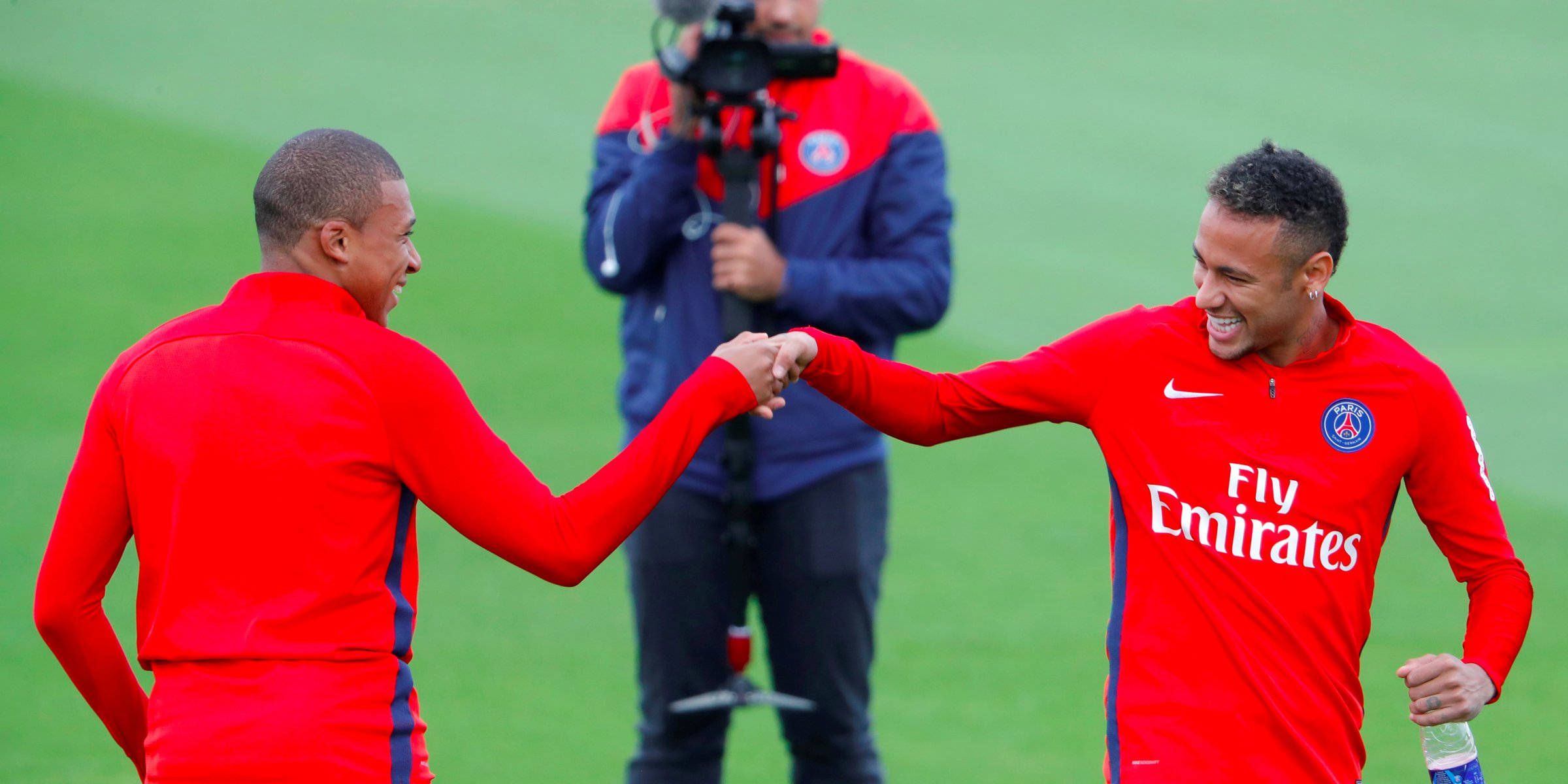 Kylian Mbappe 桌布 壁紙 — 兩名身穿紅色訓練服、面帶微笑的足球員在綠色球場上握手，背景有一位攝影師。
