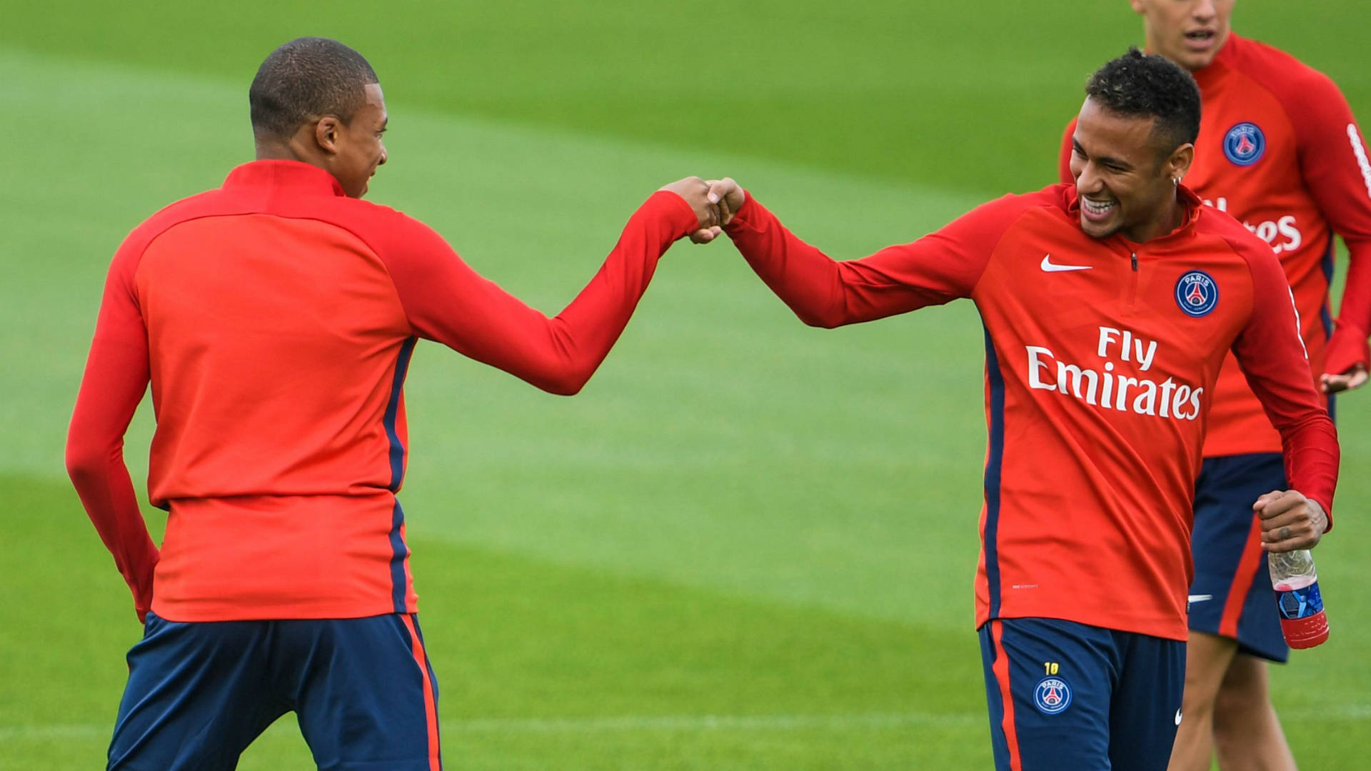 Kylian Mbappe 桌布 壁紙 — 兩名身穿紅色訓練服的足球員在陽光明媚的日子裡，在充滿活力的綠色球場上擊掌。
