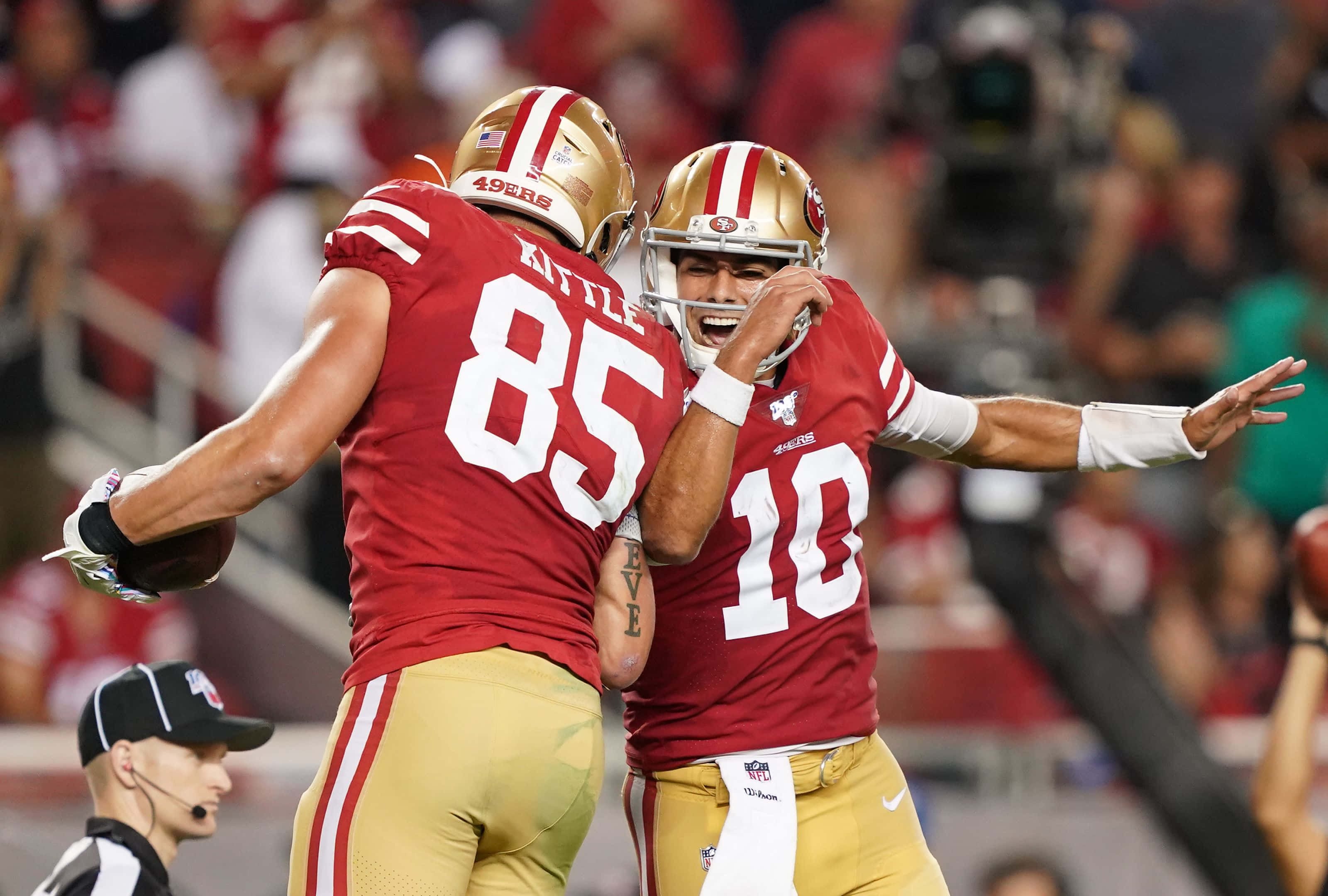 George Kittle 桌布 壁紙 — 兩名身穿紅金色球衣的舊金山四九人美式足球員在場上慶祝比賽，背景有球迷。