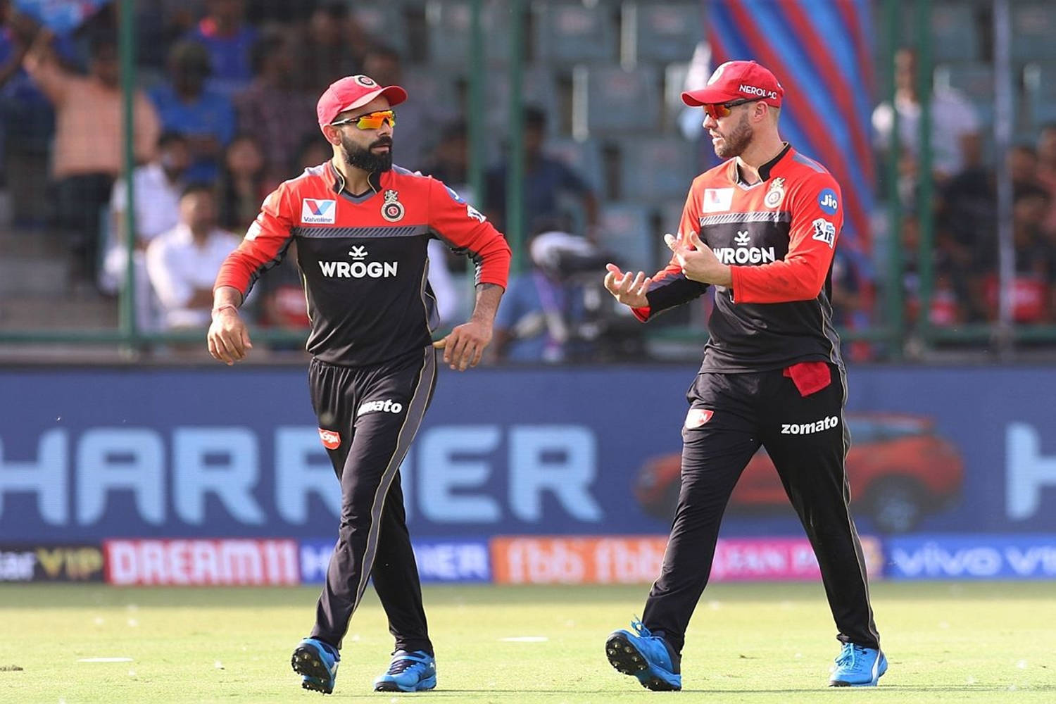 royal challengers bangalore 桌布 壁紙 — 兩名身穿紅黑球衣的板球運動員在陽光明媚的綠色球場上交談，背景是體育場觀眾。