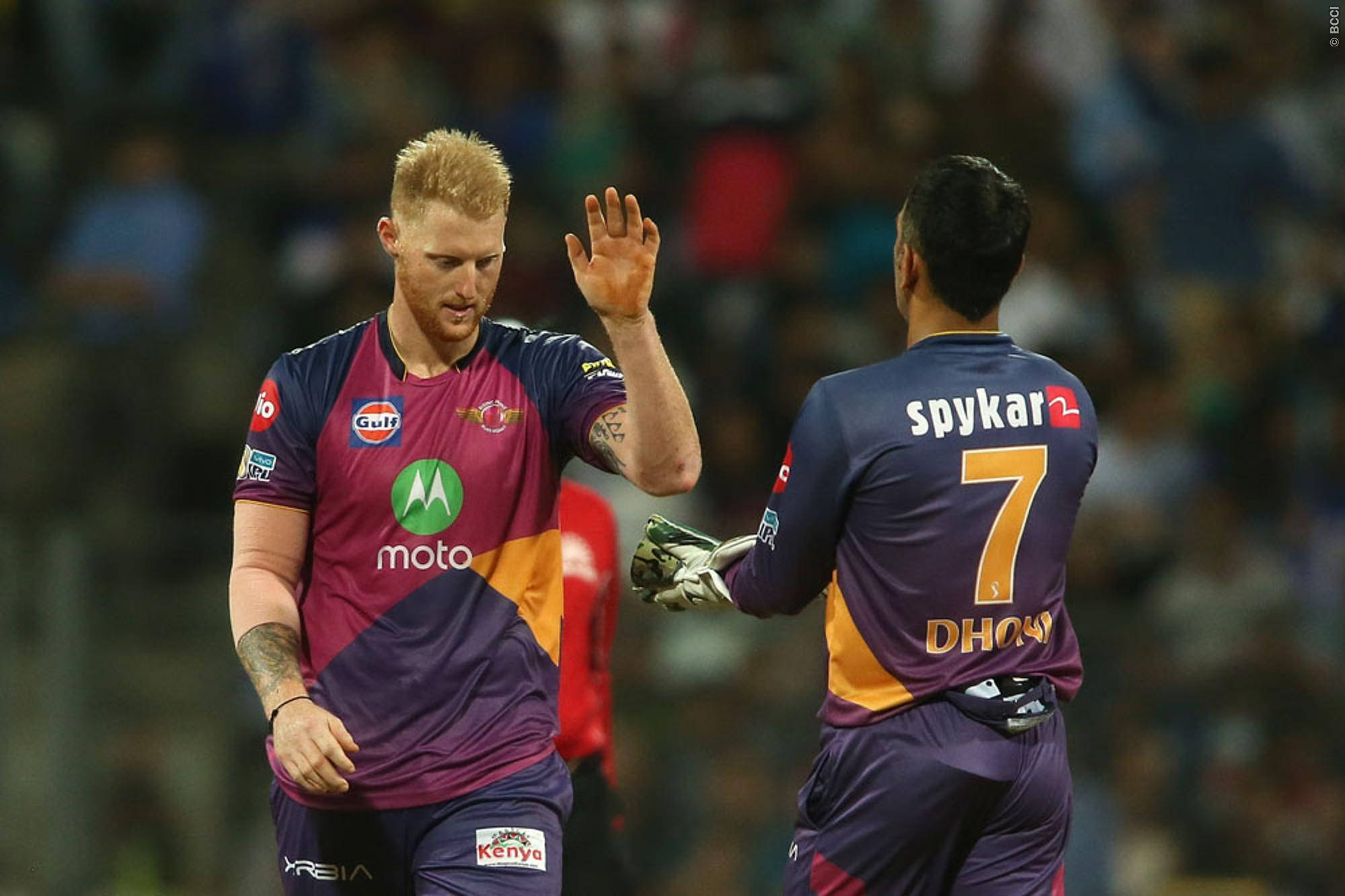 Ben Stokes 桌布 壁紙 — 兩名身穿紫色和橙色球衣的板球運動員在體育場燈光下的夜間比賽中擊掌。