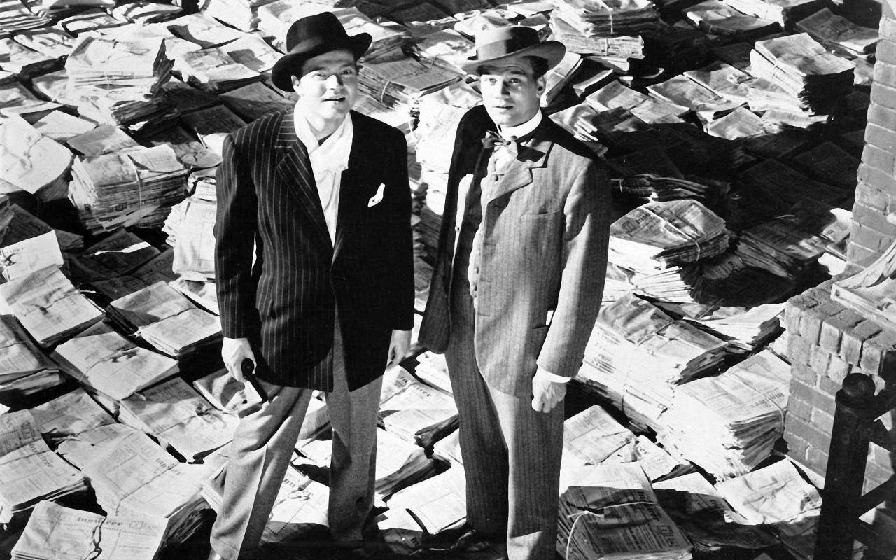Citizen Kane 桌布 壁紙 — 兩名身穿細條紋西裝和帽子的男子，在陽光明媚的城市環境中，站在高聳的報紙堆之間。