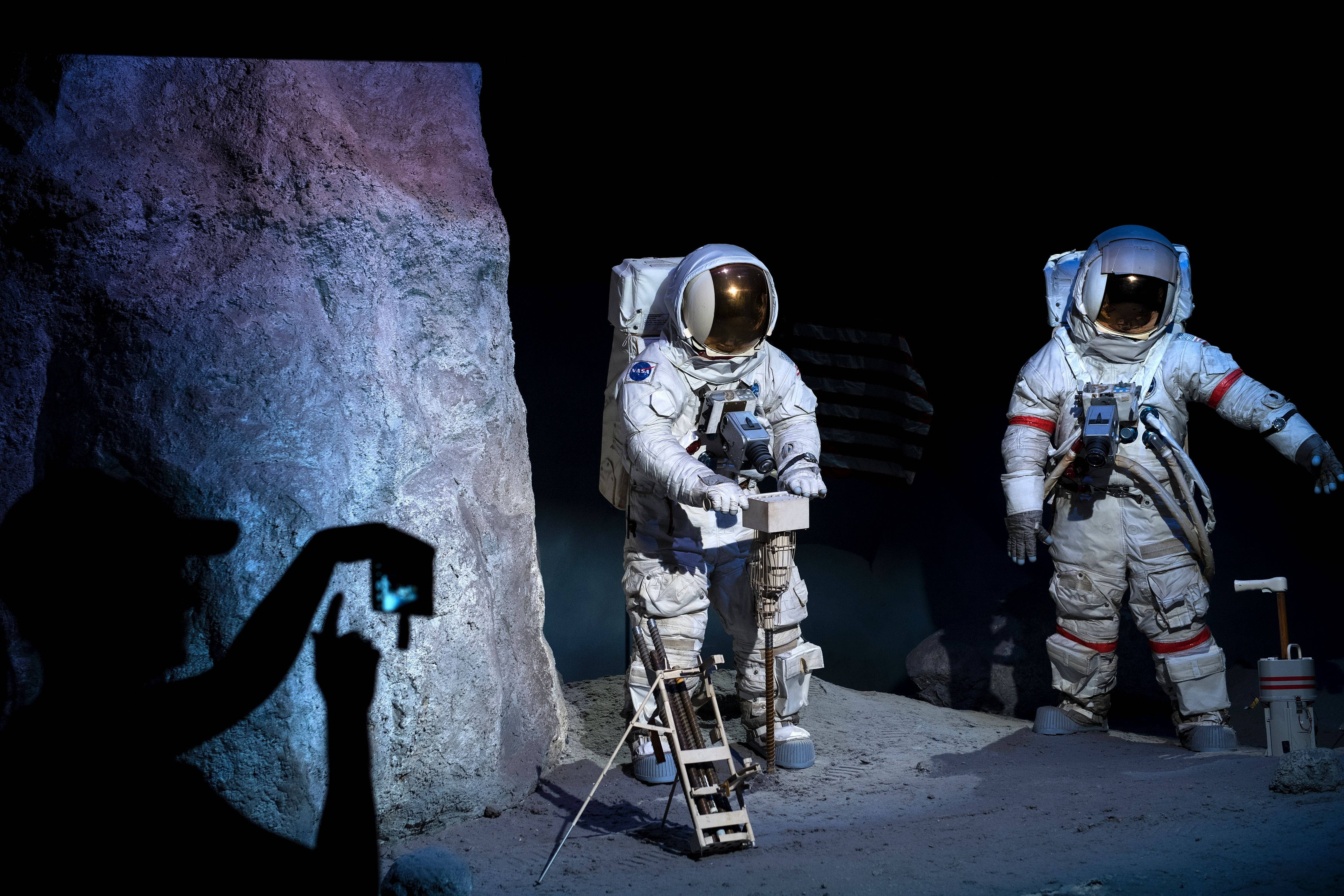 Nasa 桌布 壁紙 — 兩名身穿細節太空衣的太空人站在月球般的表面，旁邊有岩石結構，一個剪影人物正在拍攝他們。