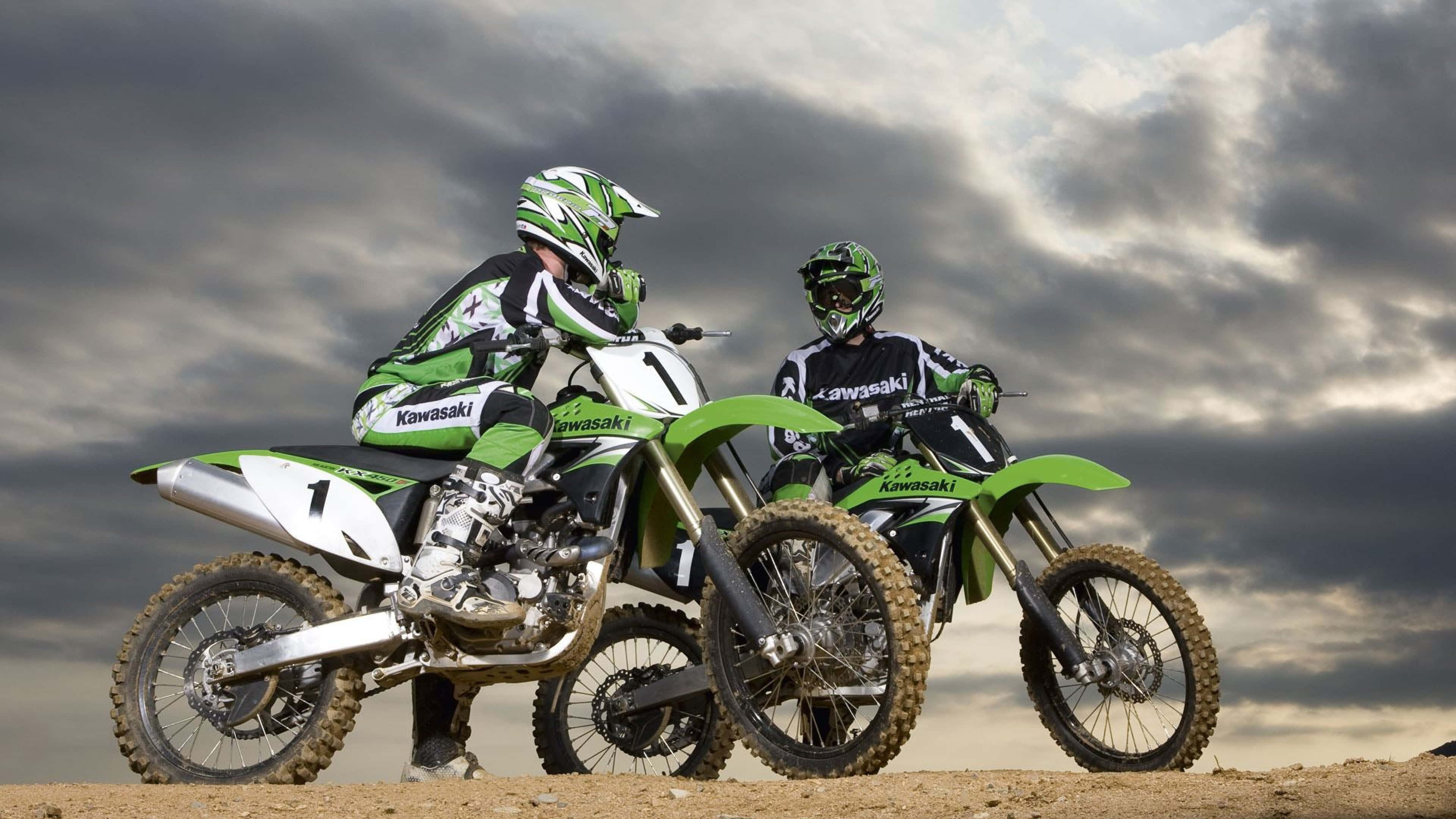 Honda Dirt Bike 桌布 壁紙 — 兩名身穿綠色和白色裝備的越野摩托車手，在戲劇性的多雲天空下，坐在他們的川崎越野車上蓄勢待發。