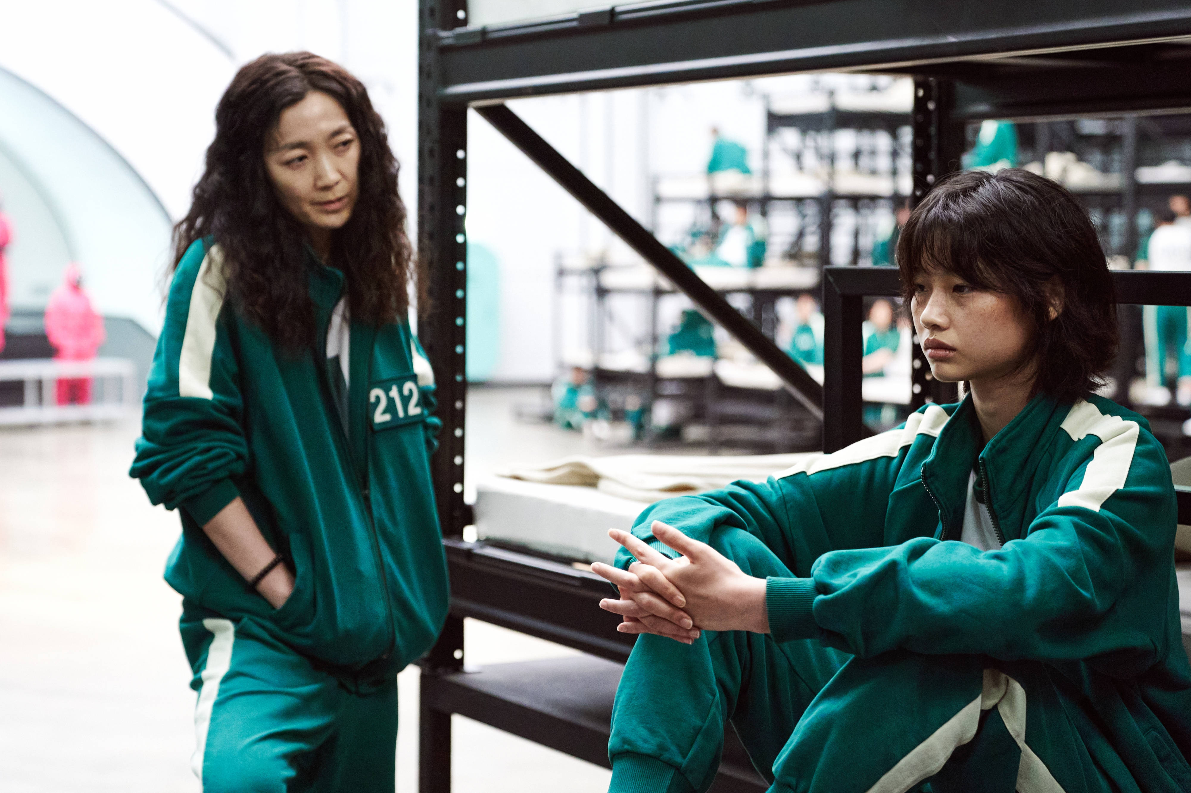 game 桌布 壁紙 — 兩名身穿綠色號碼運動服的女性坐在一個空曠的倉庫裡，背景有金屬架和其他球員。