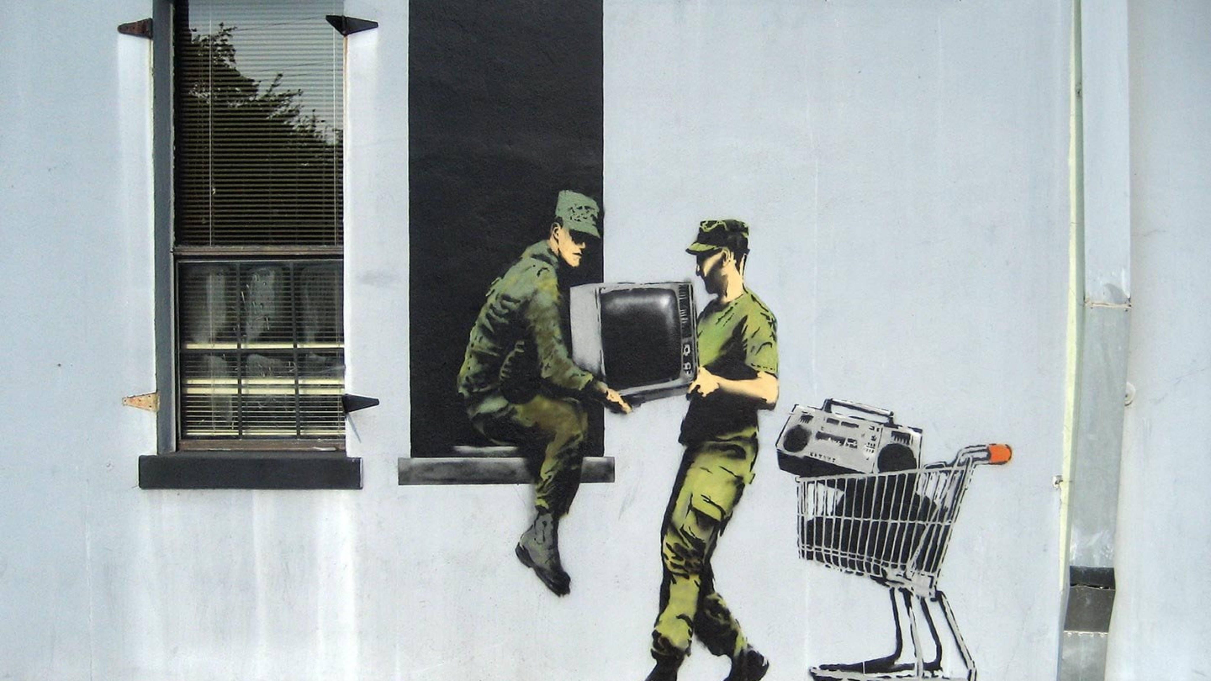 Banksy 桌布 壁紙 — 兩名身穿綠色軍裝的士兵在白色牆壁壁畫上，與購物車旁邊的復古科技（電視和手提音響）互動。