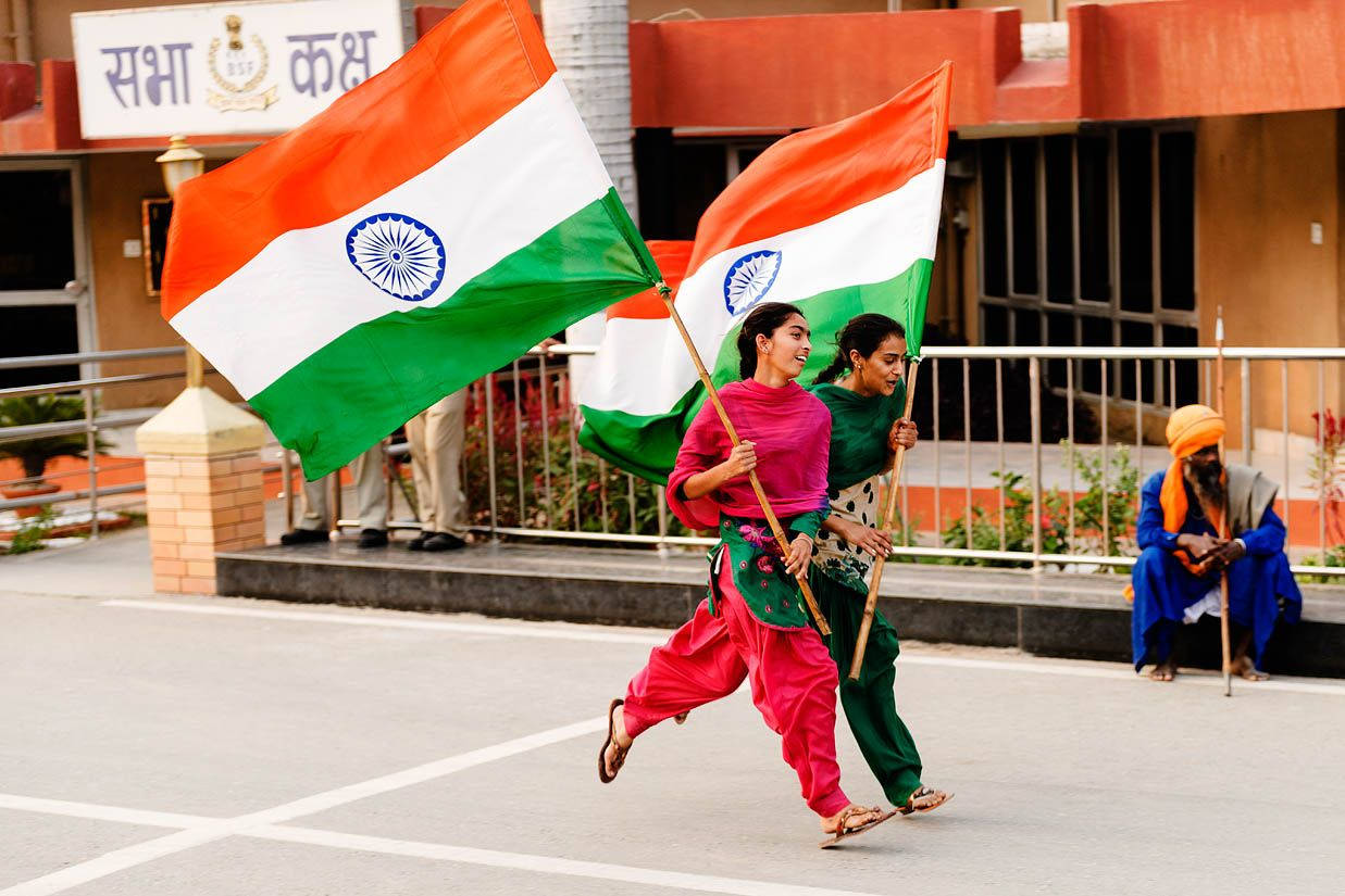 Indian 桌布 壁紙 — 兩名身穿色彩繽紛傳統服飾的年輕女性，在學校建築前開心地奔跑，揮舞著大型印度國旗。