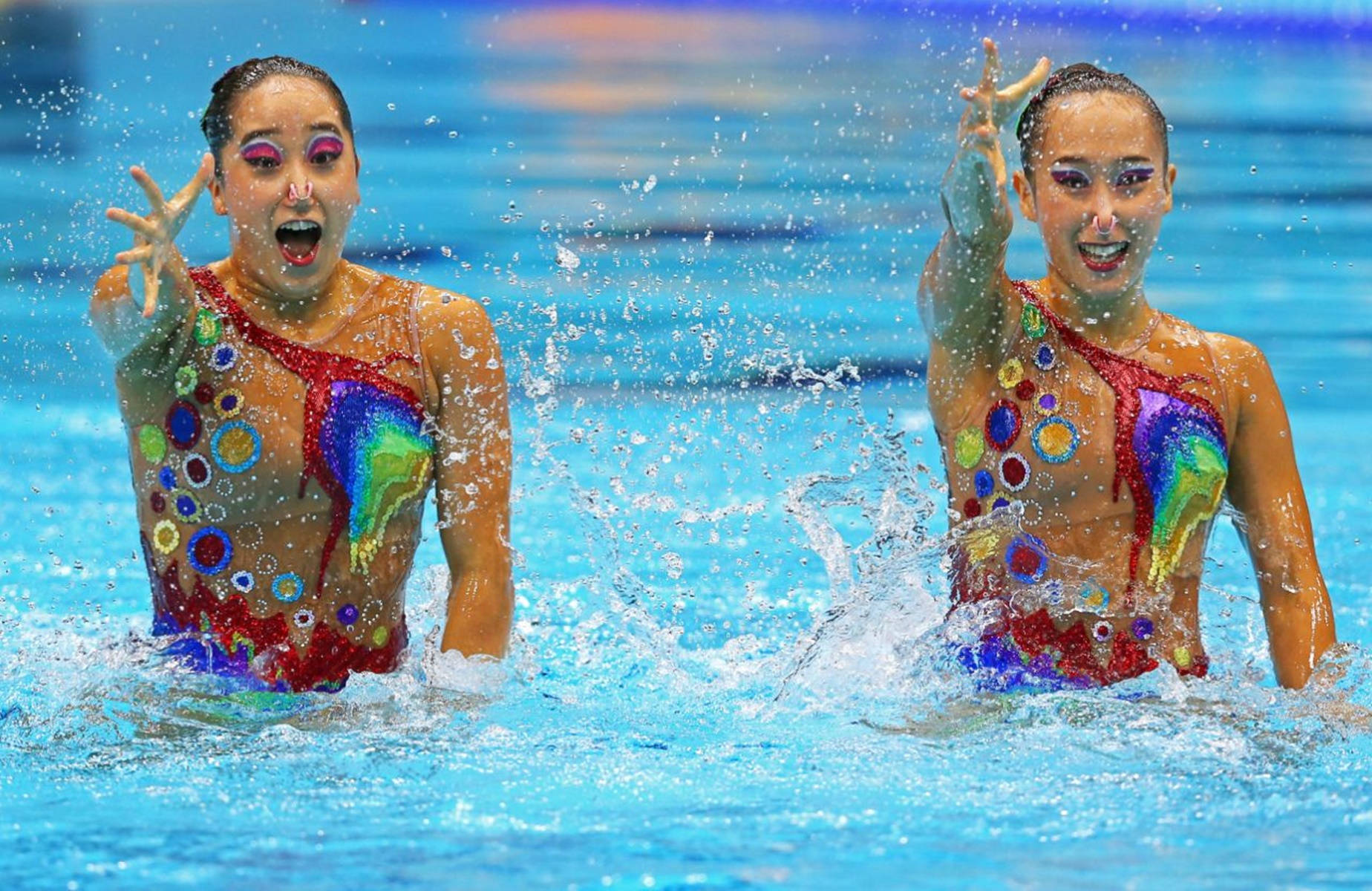 Swimming 桌布 壁紙 — 兩名身穿色彩繽紛服裝的韻律泳選手在明亮的藍色泳池中表演時歡快地濺起水花。
